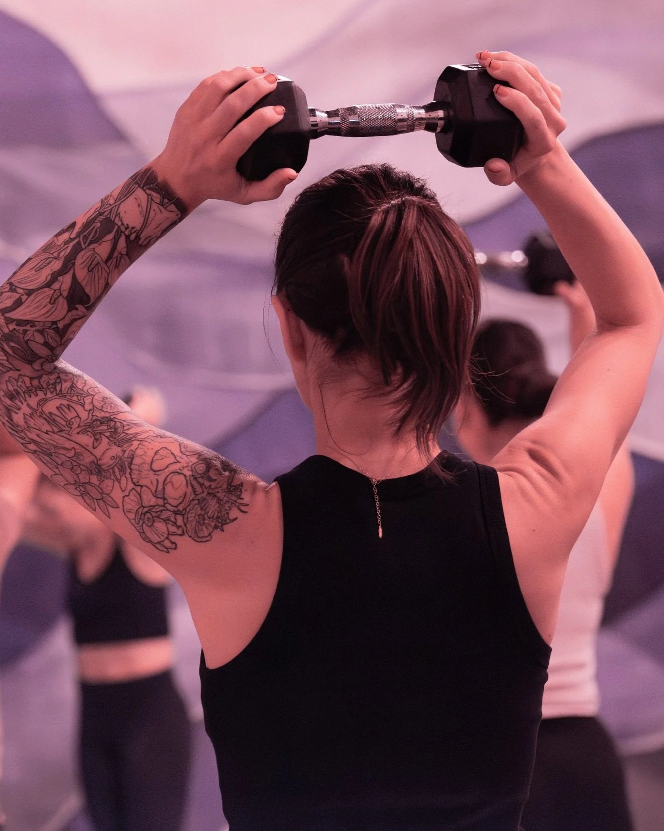 Tattooed woman lifting a dumbbell overhead in a pink-lit gym
