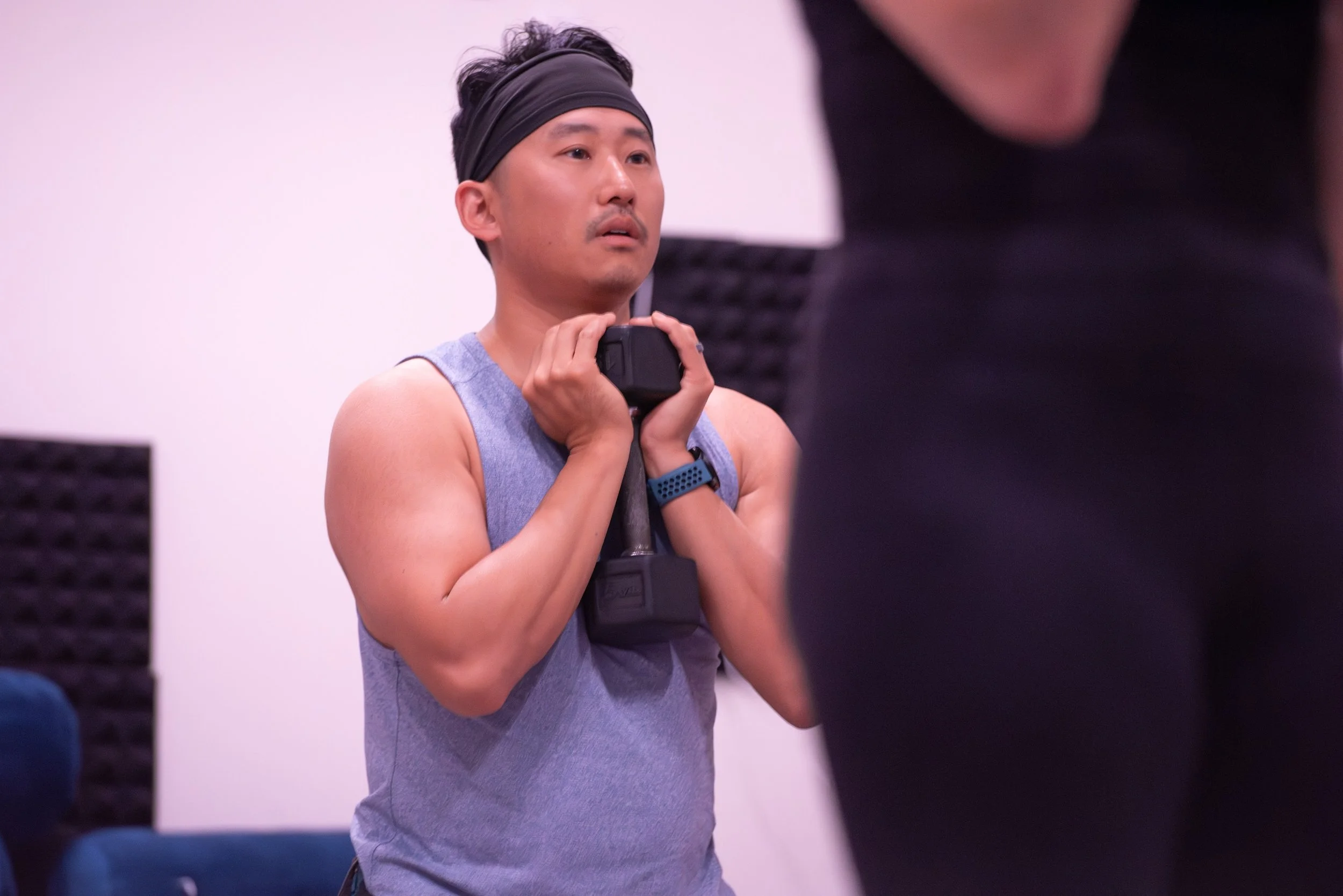 Man in a gray tank top and headband holding a black dumbbell during a workout