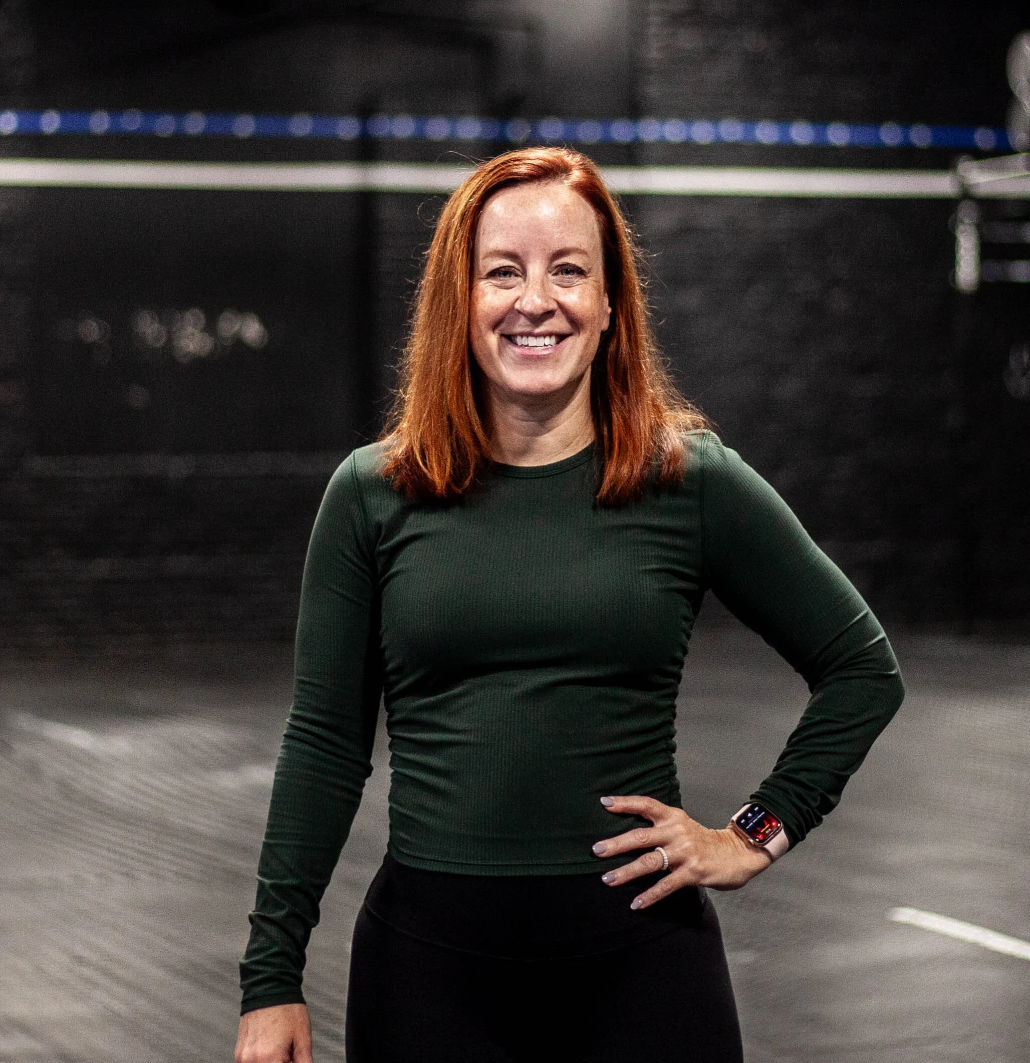 A woman with long red hair wearing a dark green long-sleeve shirt, smiling, with her right hand on her hip, in an indoor gym or workout space.
