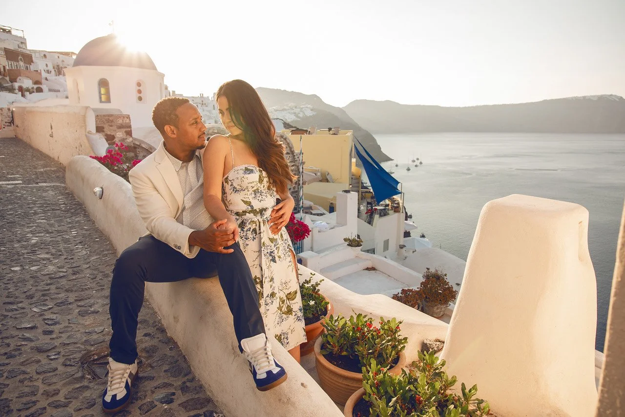 A couple sitting on a stone wall in Santorini, Greece, with a view of the Aegean Sea and whitewashed buildings. The man is wearing a white jacket and jeans, and the woman is in a floral dress. There are potted plants beside them, and the sun is setting.