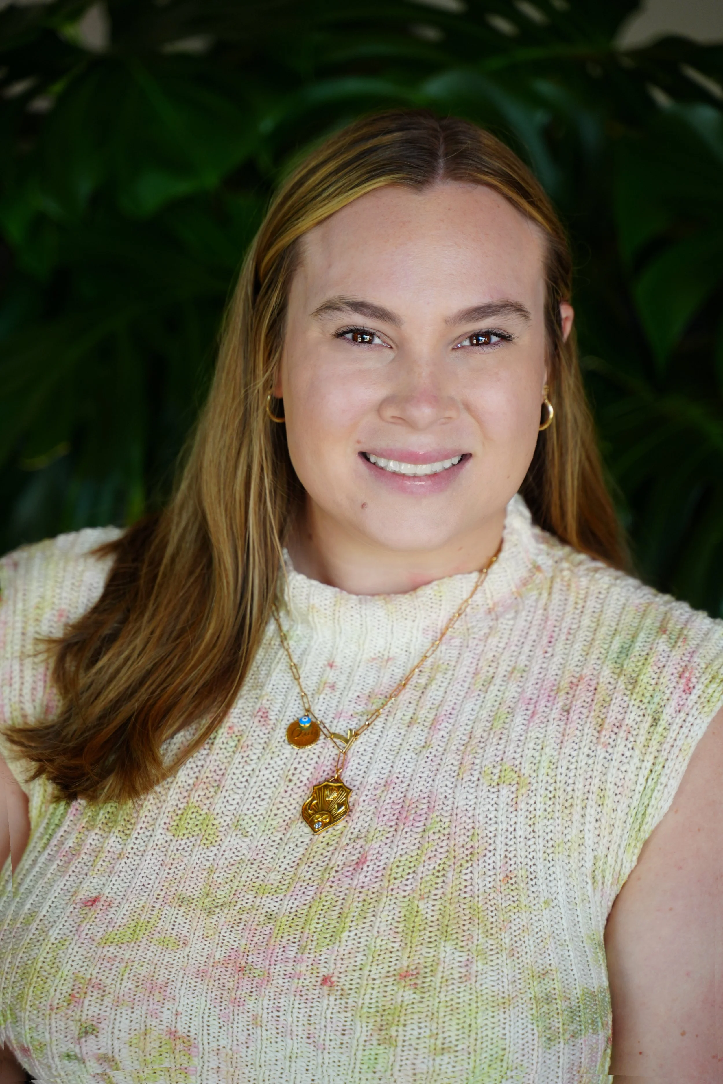 Smiling person wearing a knitted sleeveless top, gold hoop earrings, and layered necklaces, standing against a backdrop of large green leaves.