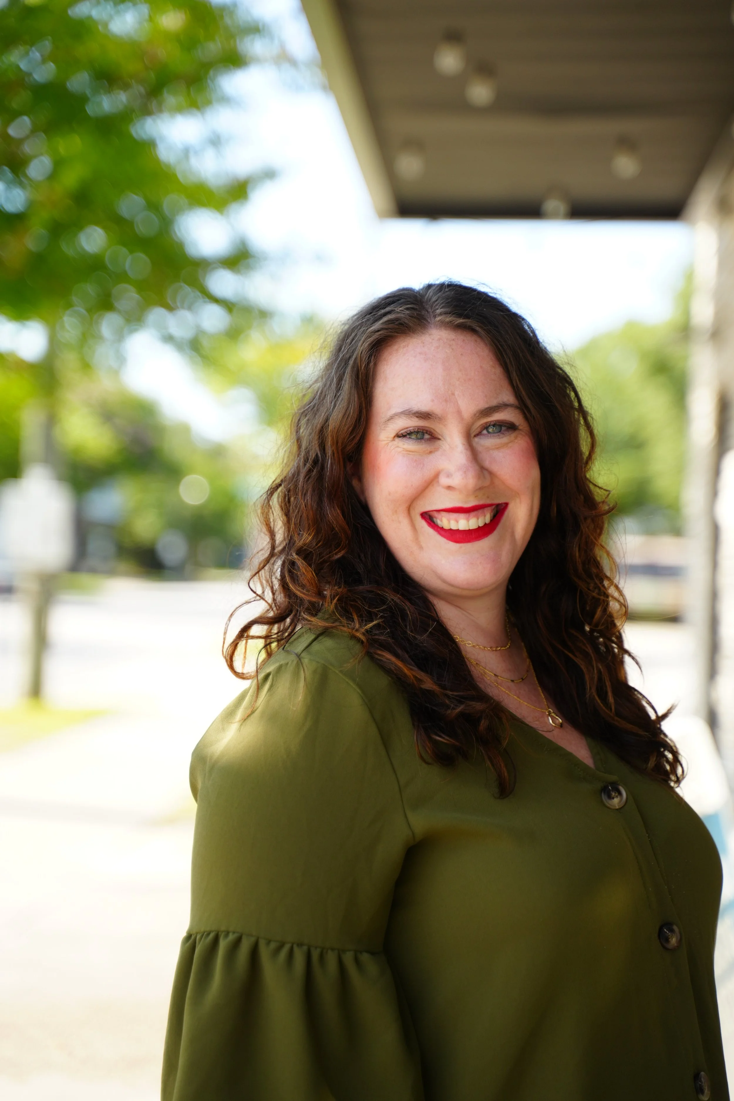 Smiling woman with curly hair wearing an olive green blouse standing outdoors.