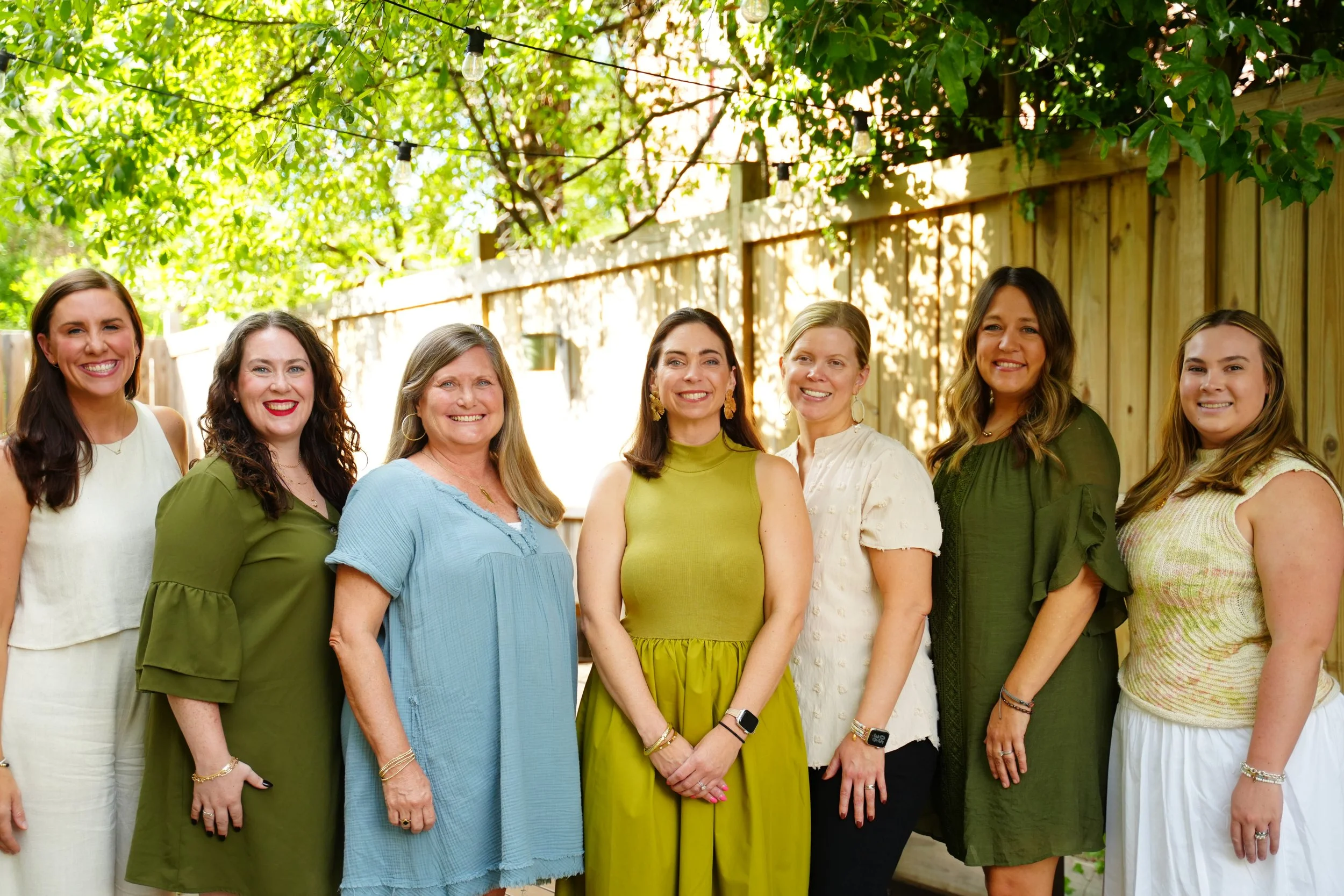Group of seven women smiling outdoors with green trees and a wooden fence in the background, dressed in casual summer clothing.