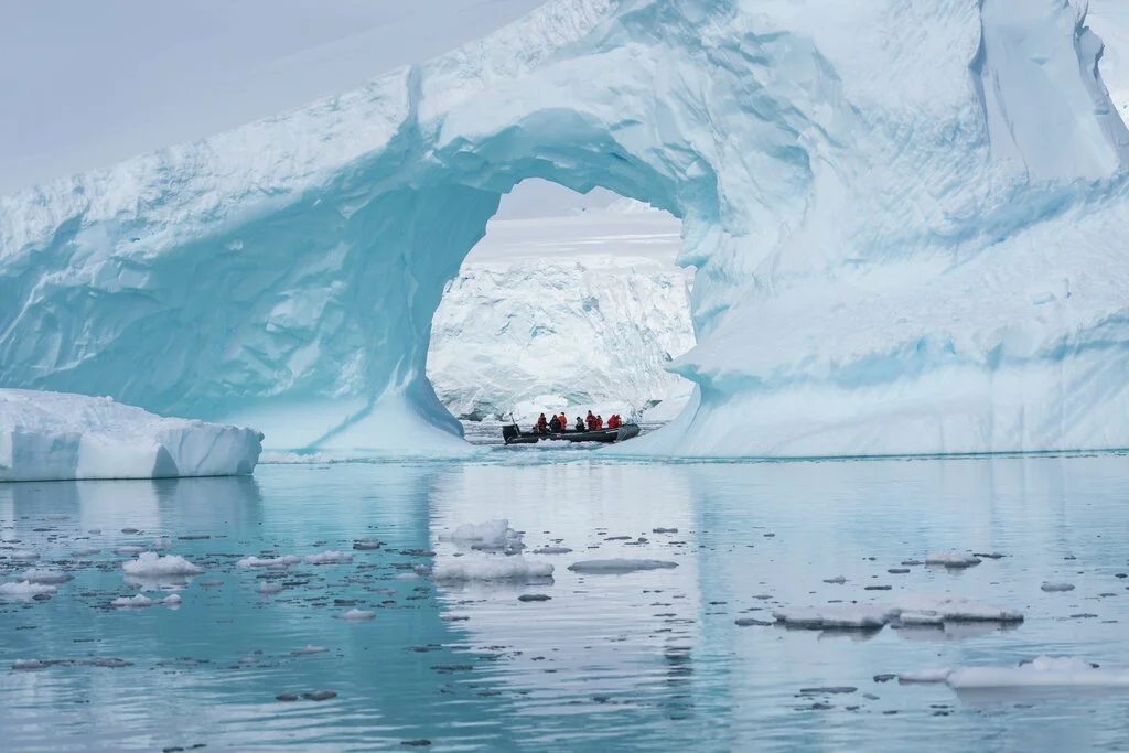 A group of people in a small boat approaching a large iceberg with a hole in it on an icy body of water.