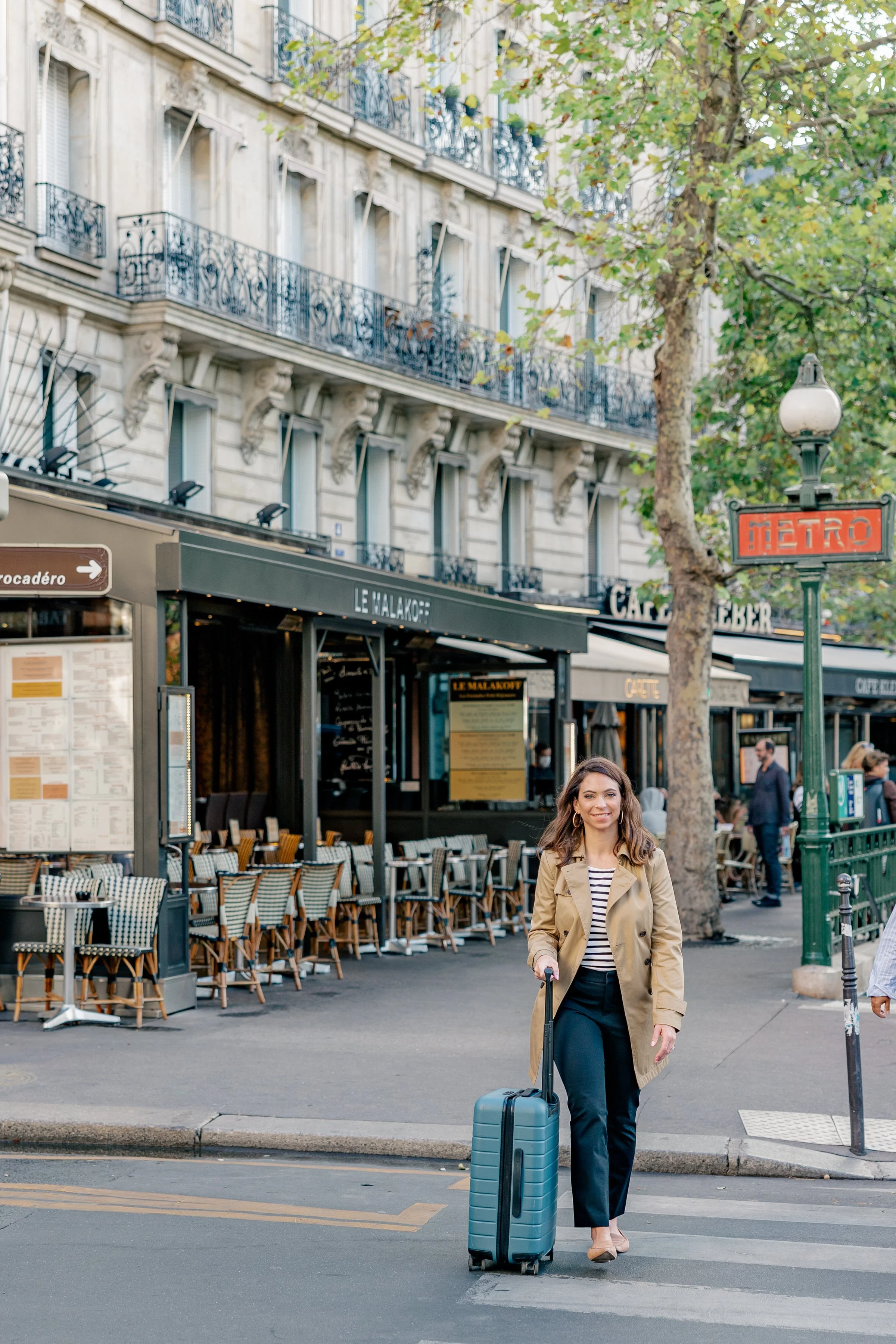 A woman walking with a suitcase on a Paris street, near a café and a Metro sign, with a classic Parisian building in the background.