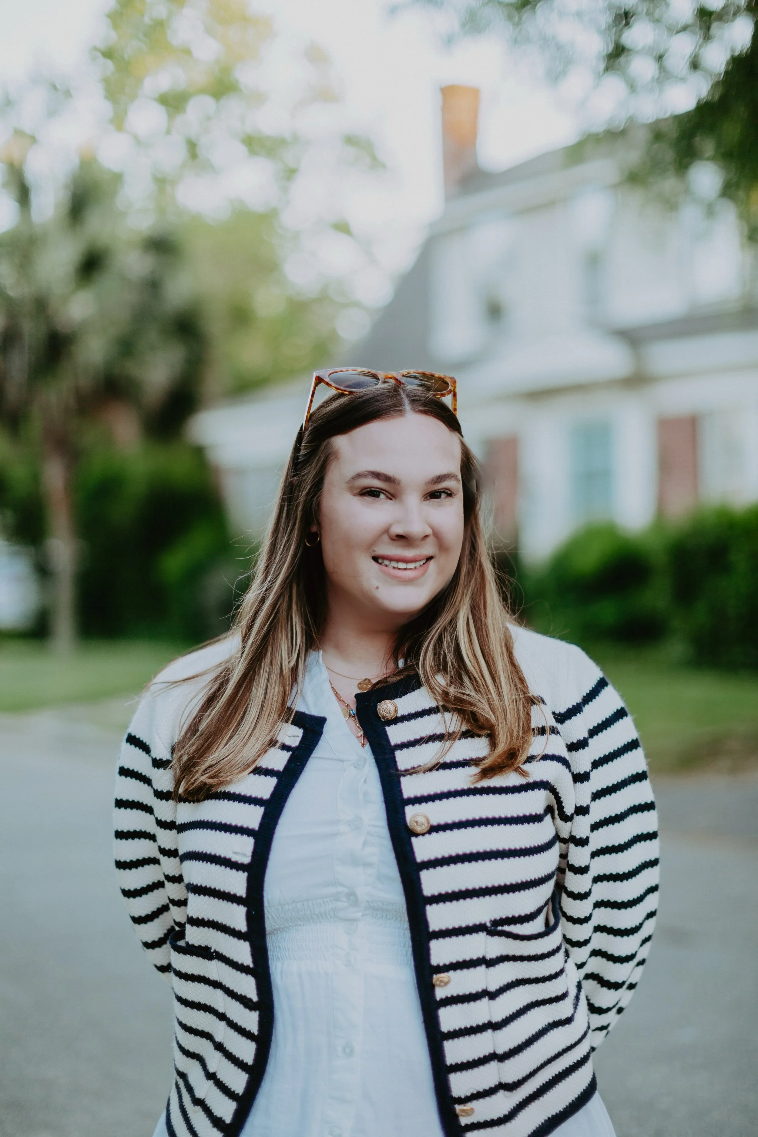 Person outdoors smiling, wearing a striped sweater with sunglasses on head, trees and house in background.