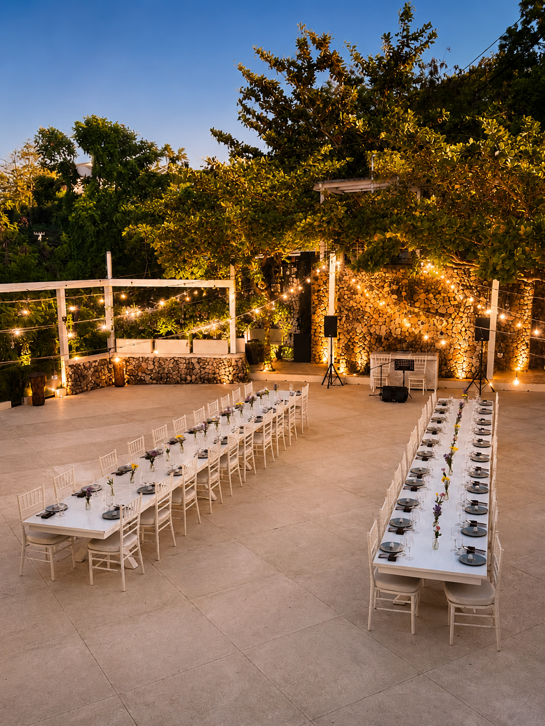 Outdoor event space with long dining tables decorated with small flower arrangements, string lights, forest background, and a blue sky at dusk.