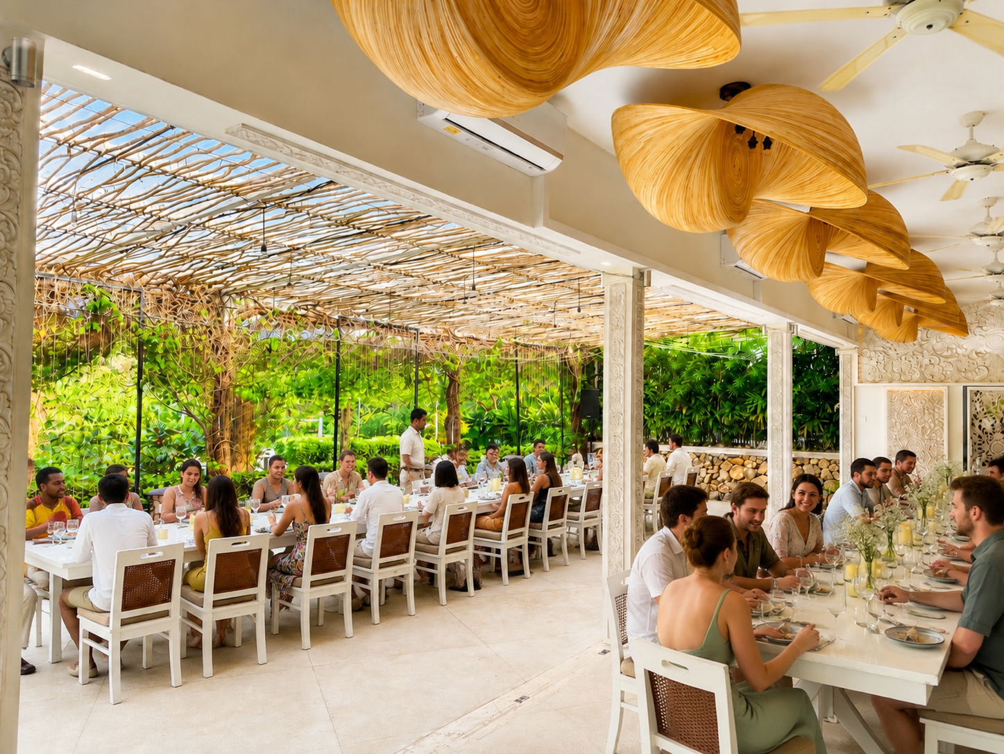 A lively restaurant with many people dining at long white tables. The restaurant has a natural, tropical decor with a lush green garden view through glass walls and a ceiling adorned with large wooden leaf-shaped sculptures.
