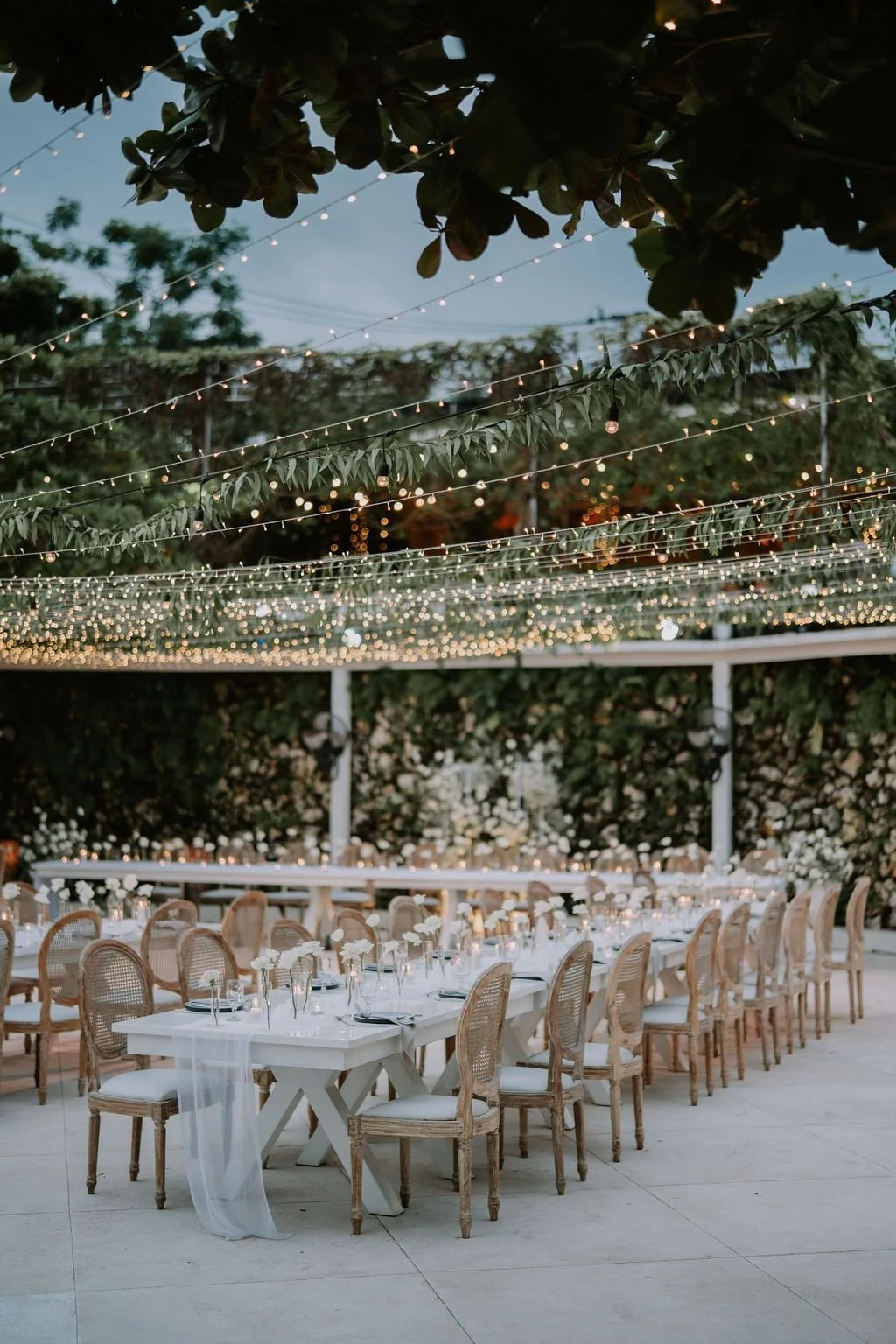 Elegant outdoor dining setup with long white tables, chairs, and floral centerpieces, under string lights and greenery canopy at dusk.