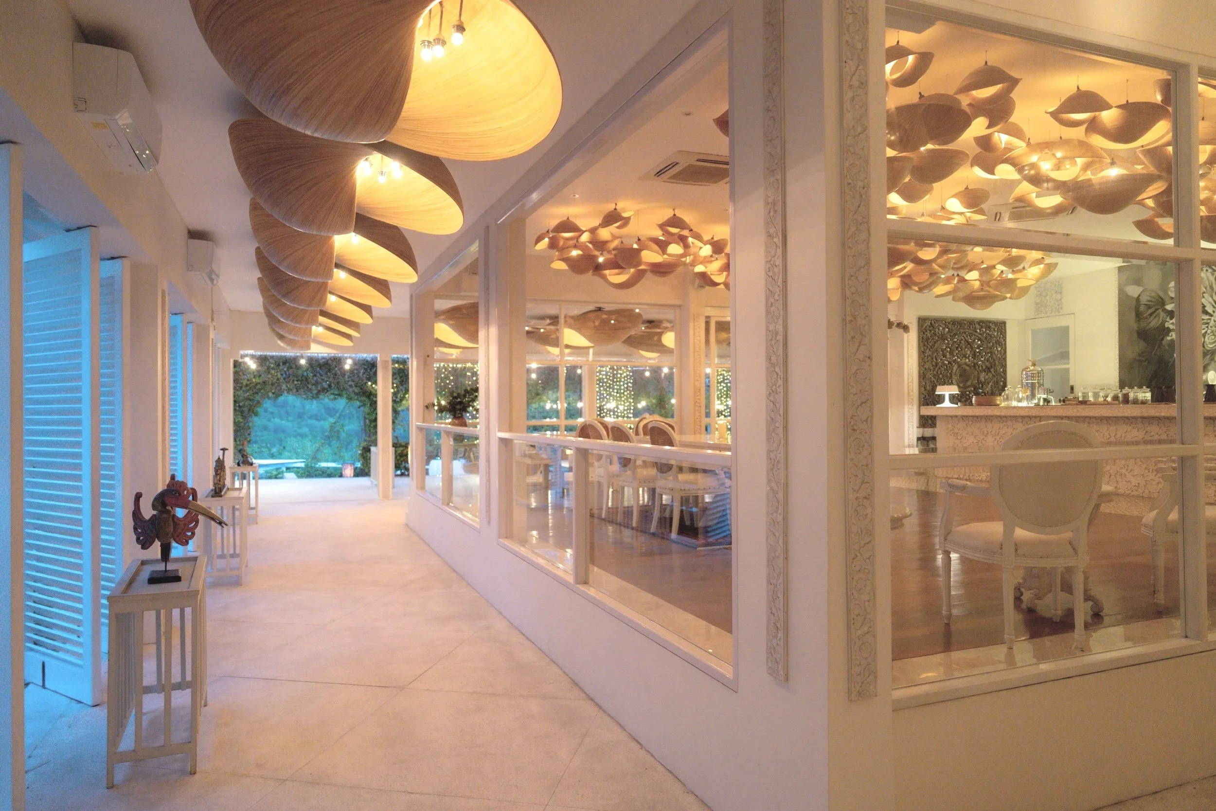 A bright indoor dining area with wooden pendant lights on the ceiling, white walls, and large windows showing an outdoor patio with greenery and string lights.