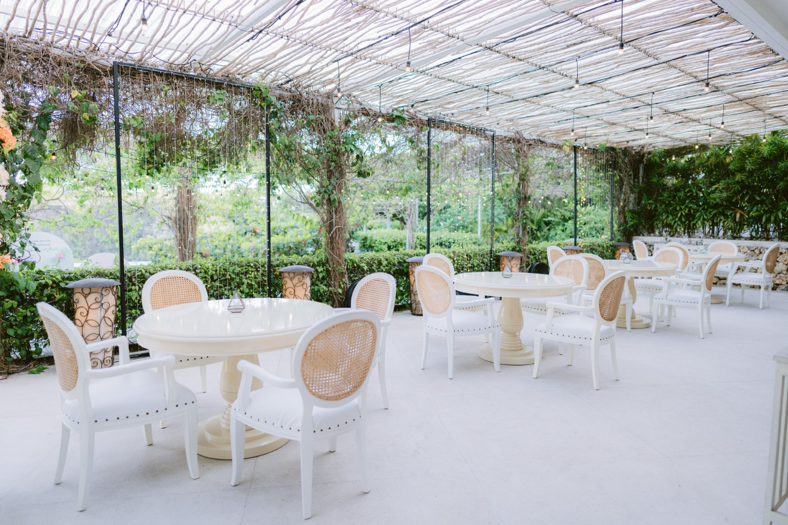 An outdoor patio with white round tables and chairs, decorated with small vases, under a pergola with a bamboo roof, surrounded by greenery and plants.
