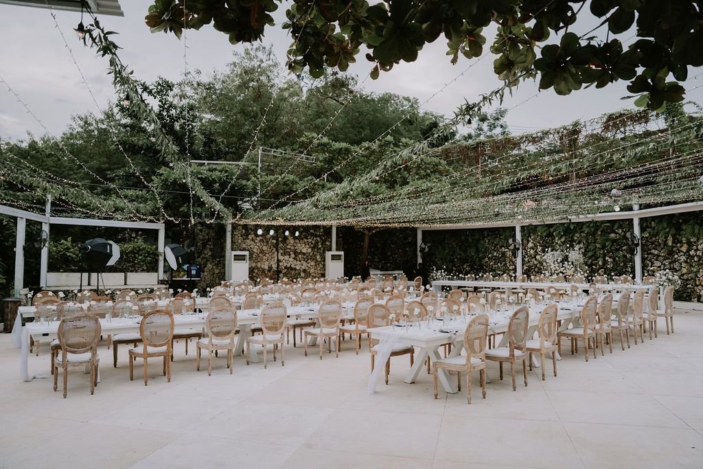 An outdoor event space decorated with string lights, set up with long dining tables and chairs for a celebration or party.
