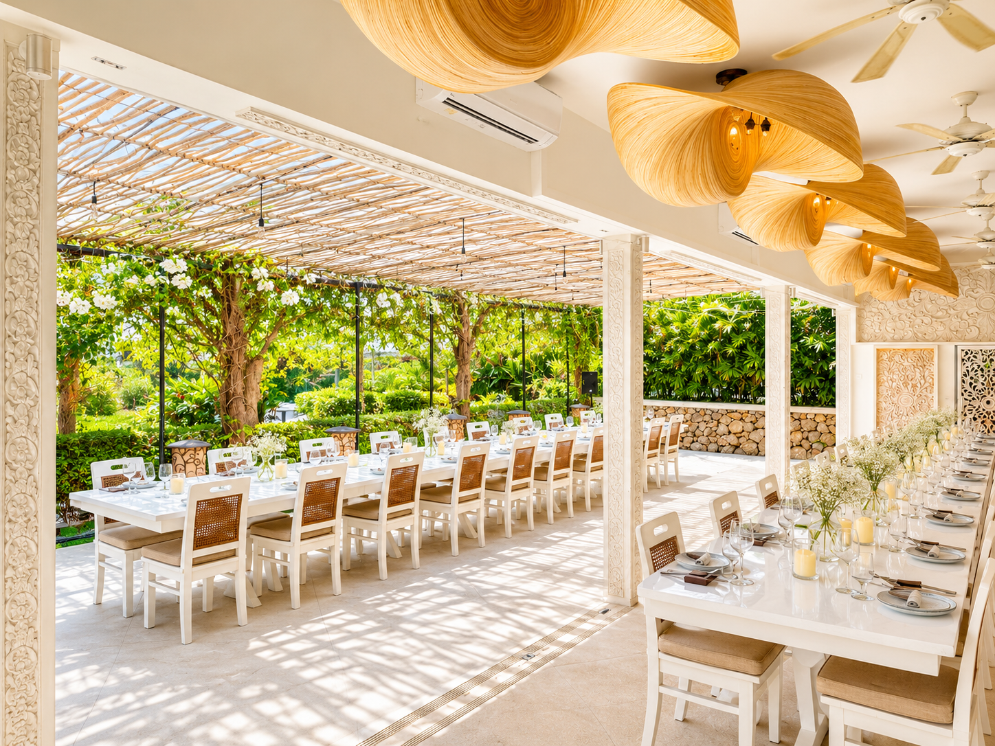 Elegant outdoor dining area with long white tables set with plates, glasses, cutlery, and floral centerpieces, surrounded by greenery and trees, with wooden ceiling fixtures and decorative columns.