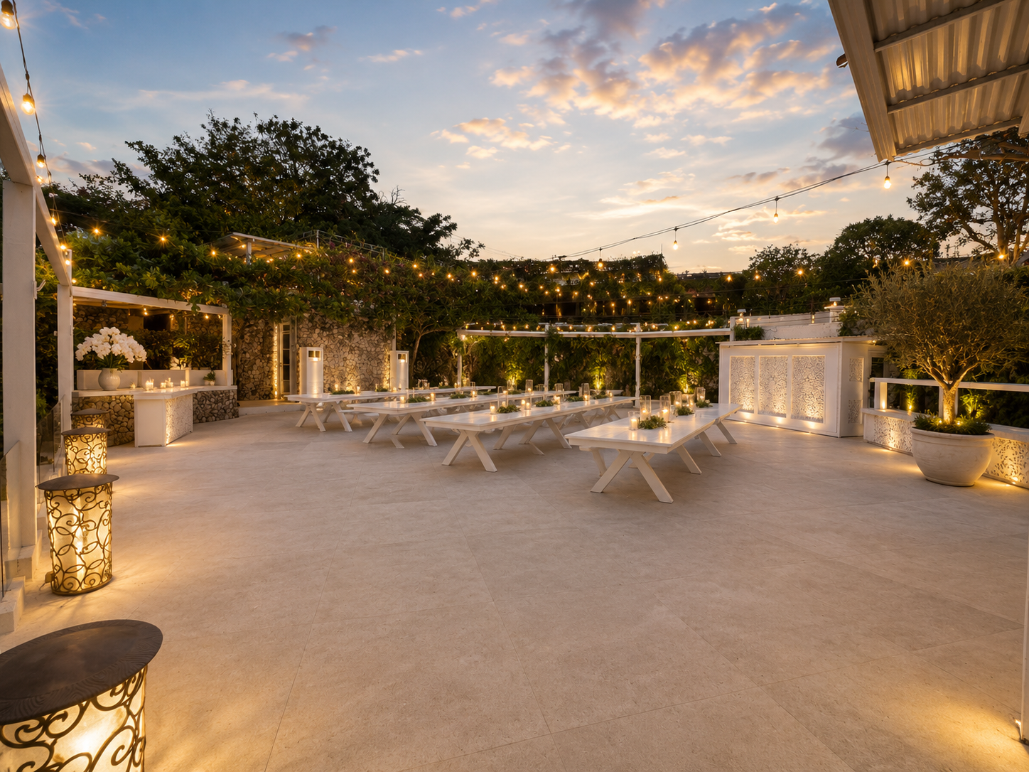 An outdoor terrace decorated for an evening event with string lights, multiple white tables, candles, potted plants, and a partly cloudy sky in the background.