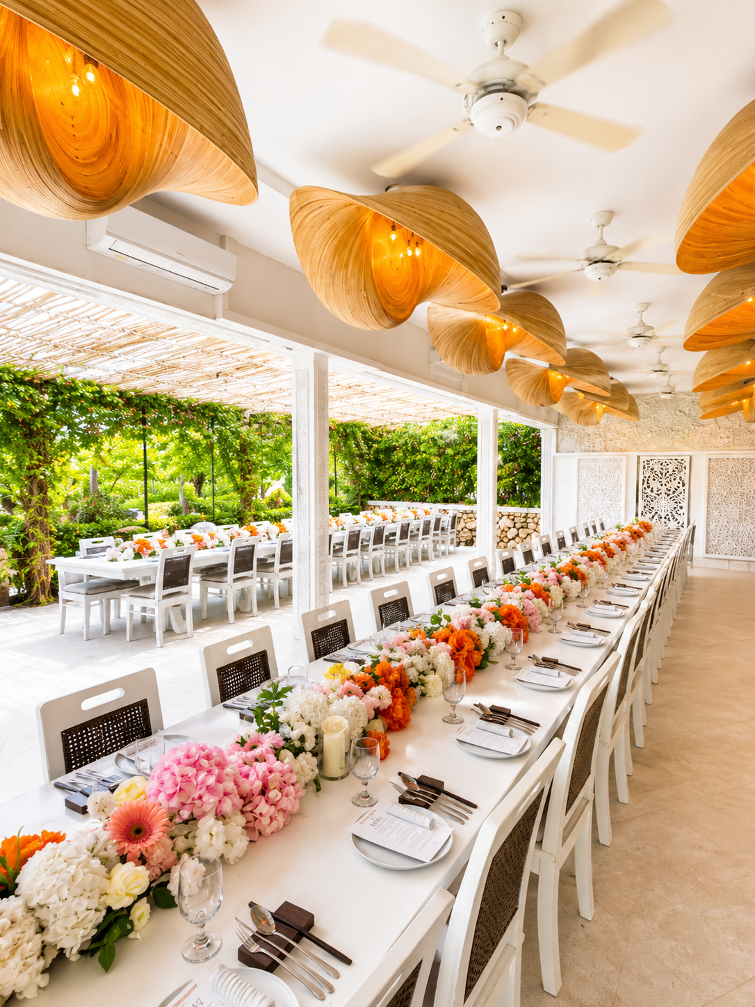 Long indoor dining table decorated with pink, white, and orange flowers, set for a formal event with place settings, in a bright room with outdoor view of greenery and ceiling fans.