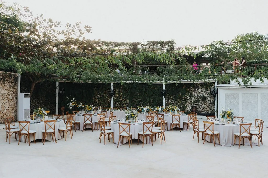 An outdoor event setup with multiple round tables covered in white tablecloths, each decorated with floral centerpieces, surrounded by wooden chairs with white cushions. There are strings of lights and people on an upper deck area in the background, 
