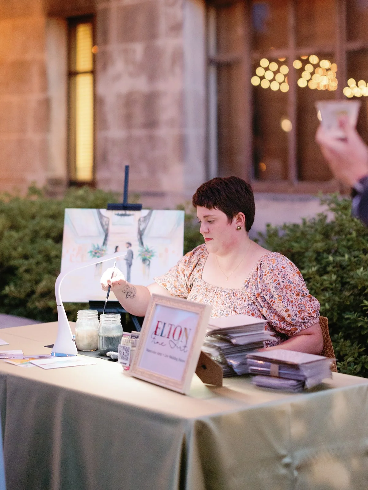 Photo by Rach Decker Photo. A live painter sits behind a table with stacks of paper and watercolor supplies, and a ceremony painting in the background.
