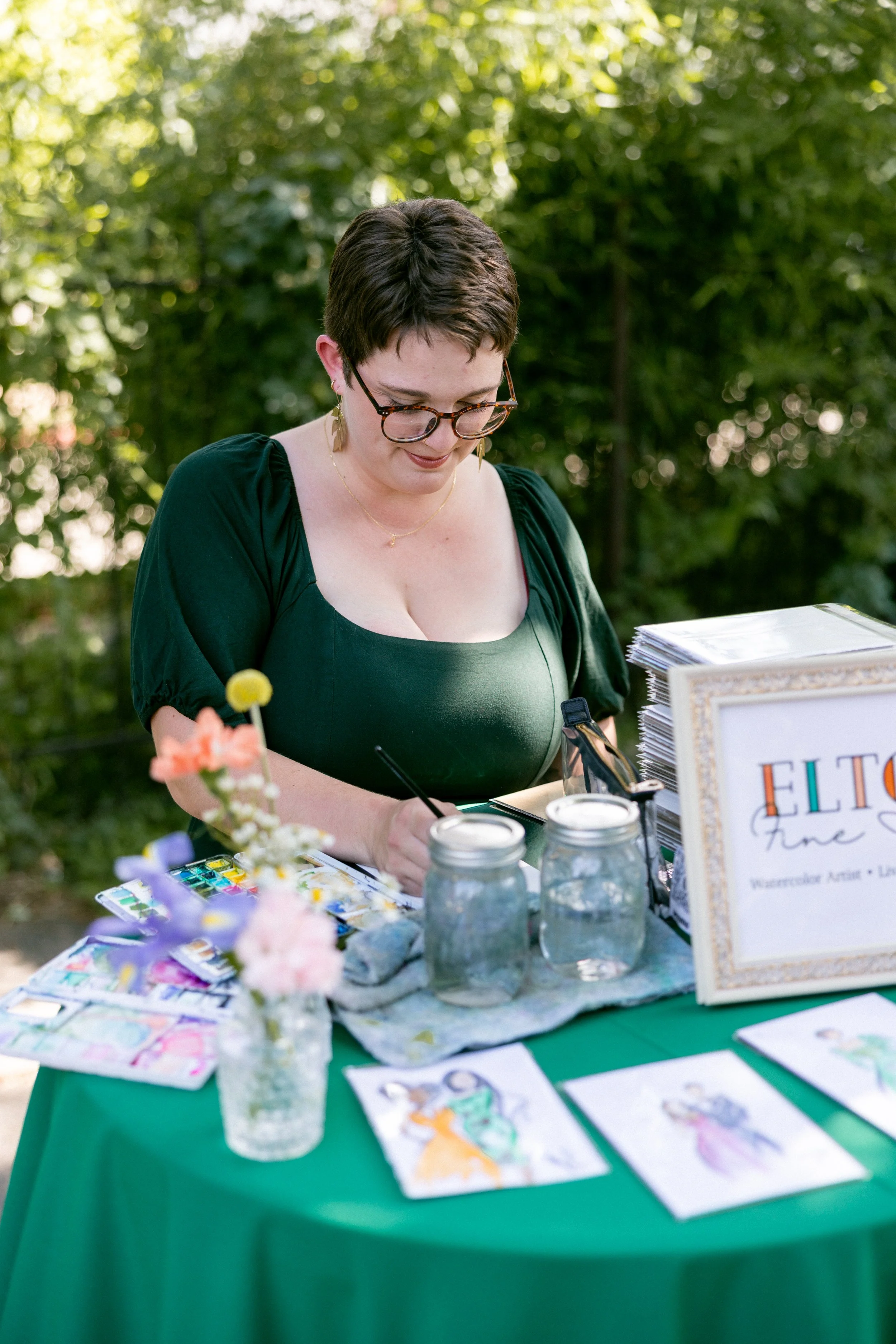 Photo by Jordan Clark Photography. Artist is working at a table with a green table cloth topped with watercolor painting supplies and a vase of flowers.