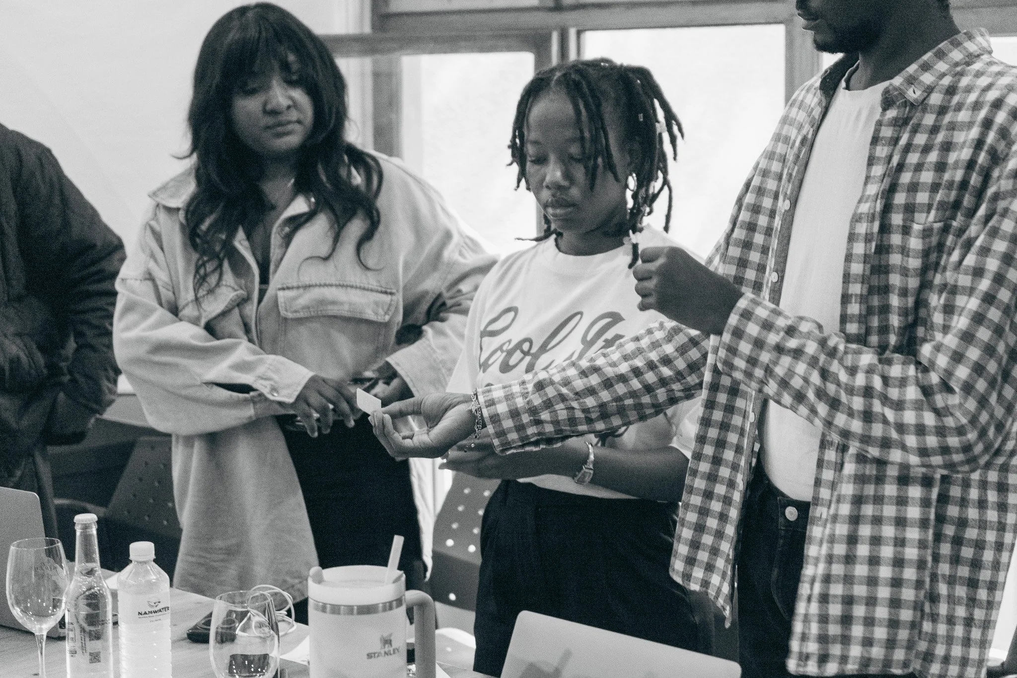 A group of three people in a meeting room, with one person handing a piece of paper to another. The table has water bottles, glasses, and a notebook.