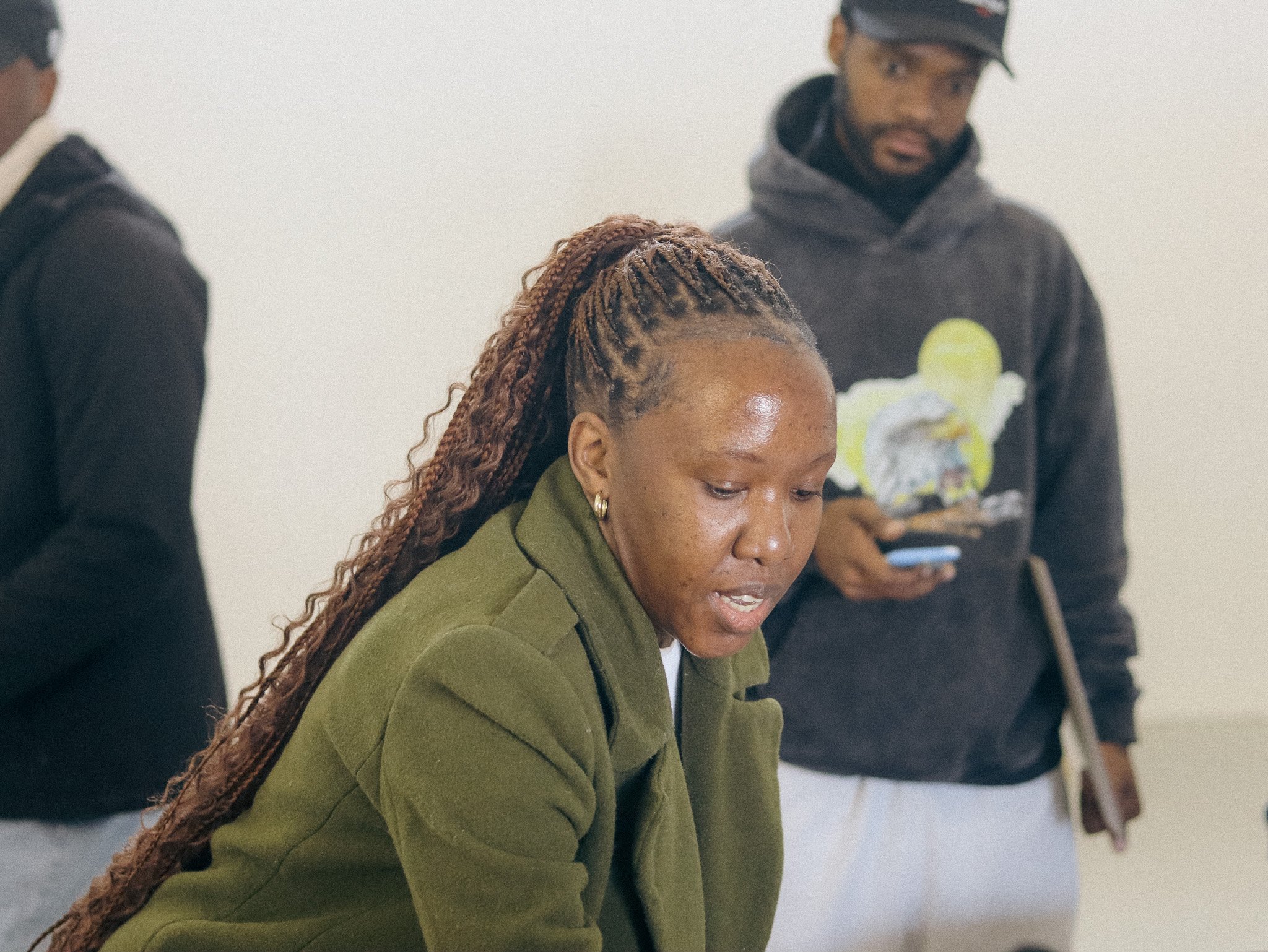 A woman with long braided hair wearing a green jacket looks down. Two men are in the background, one holding a phone and another partially visible.