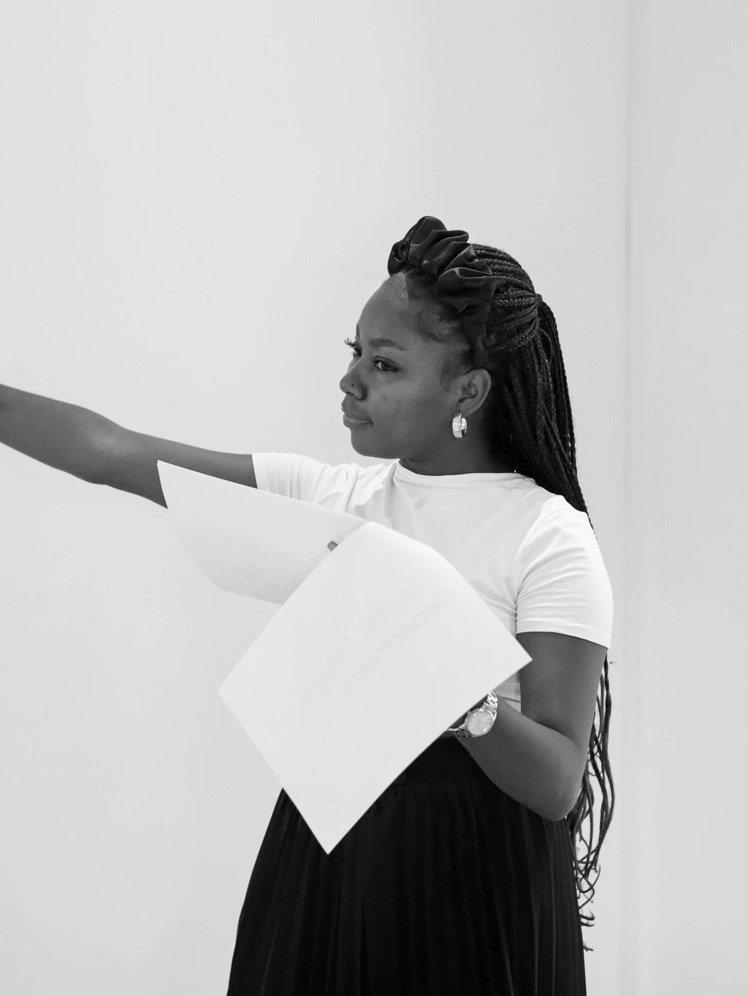 A woman with long braided hair and earrings, wearing a white t-shirt and a dark pleated skirt, is holding papers and pointing.