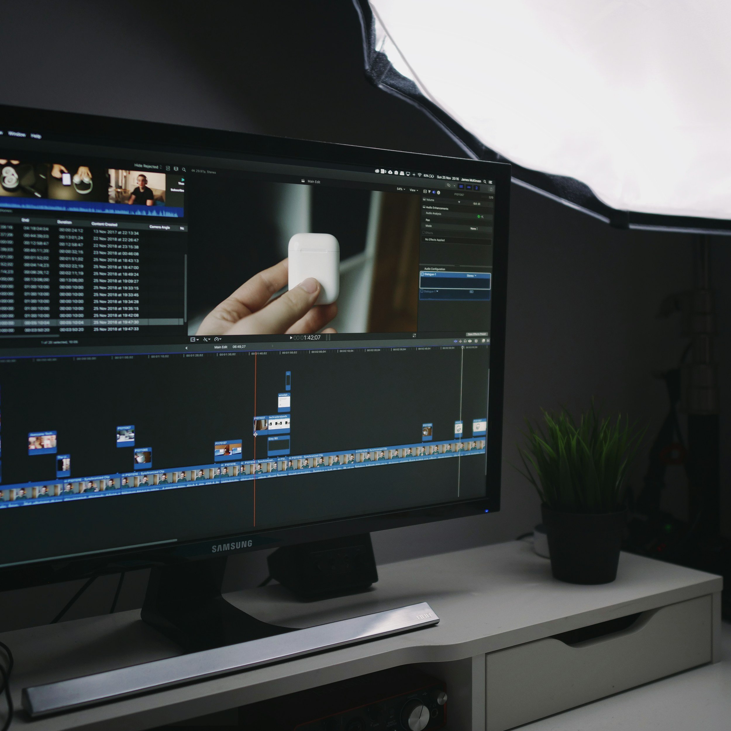 Computer monitor on a white desk displaying a video editing program with multiple video clips and editing tools visible. The monitor is black with a thin bezel, and the desk is white. A person's hand is holding a white wireless earbud case in front of the screen. The background shows a dark-colored wall and a small plant to the right.