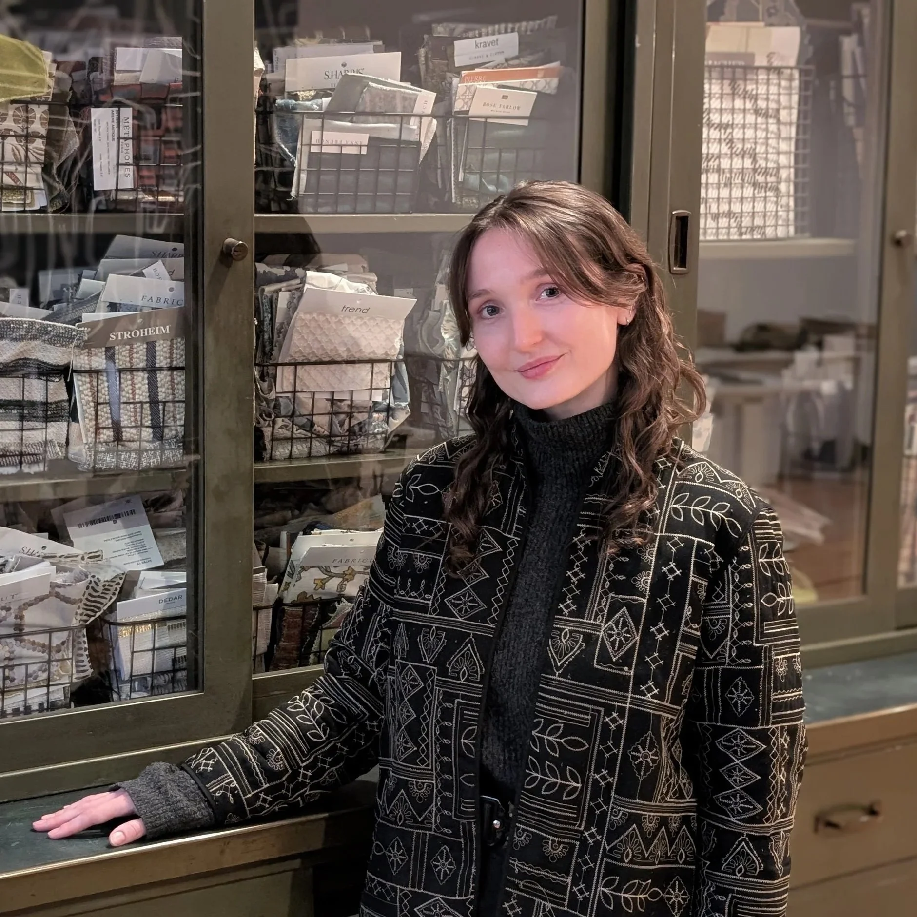 A young woman with wavy brown hair smiling and standing in front of a cabinet filled with fabric swatches and sewing supplies.
