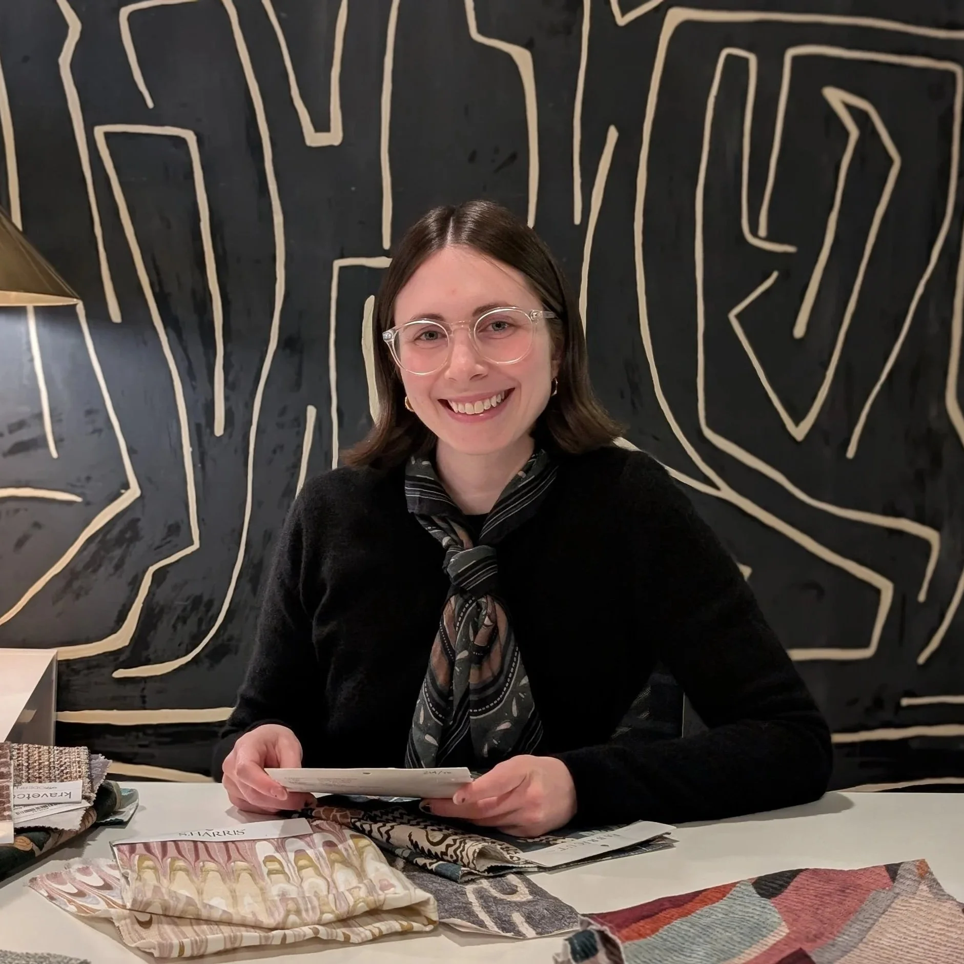 A smiling woman with short brown hair and glasses, wearing a black sweater and patterned scarf, sitting at a table with fabric samples. Behind her is a black wall with abstract beige line art.