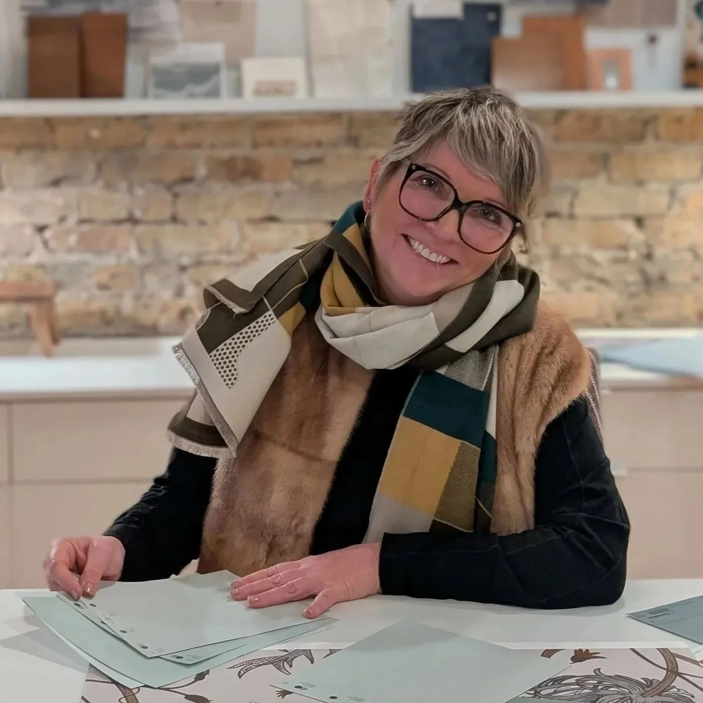 A woman with short gray hair, glasses, and a big smile sitting at a table with papers in front of her. She is wearing a multi-colored scarf, a fur vest, and a black long sleeve shirt, with a brick wall in the background.