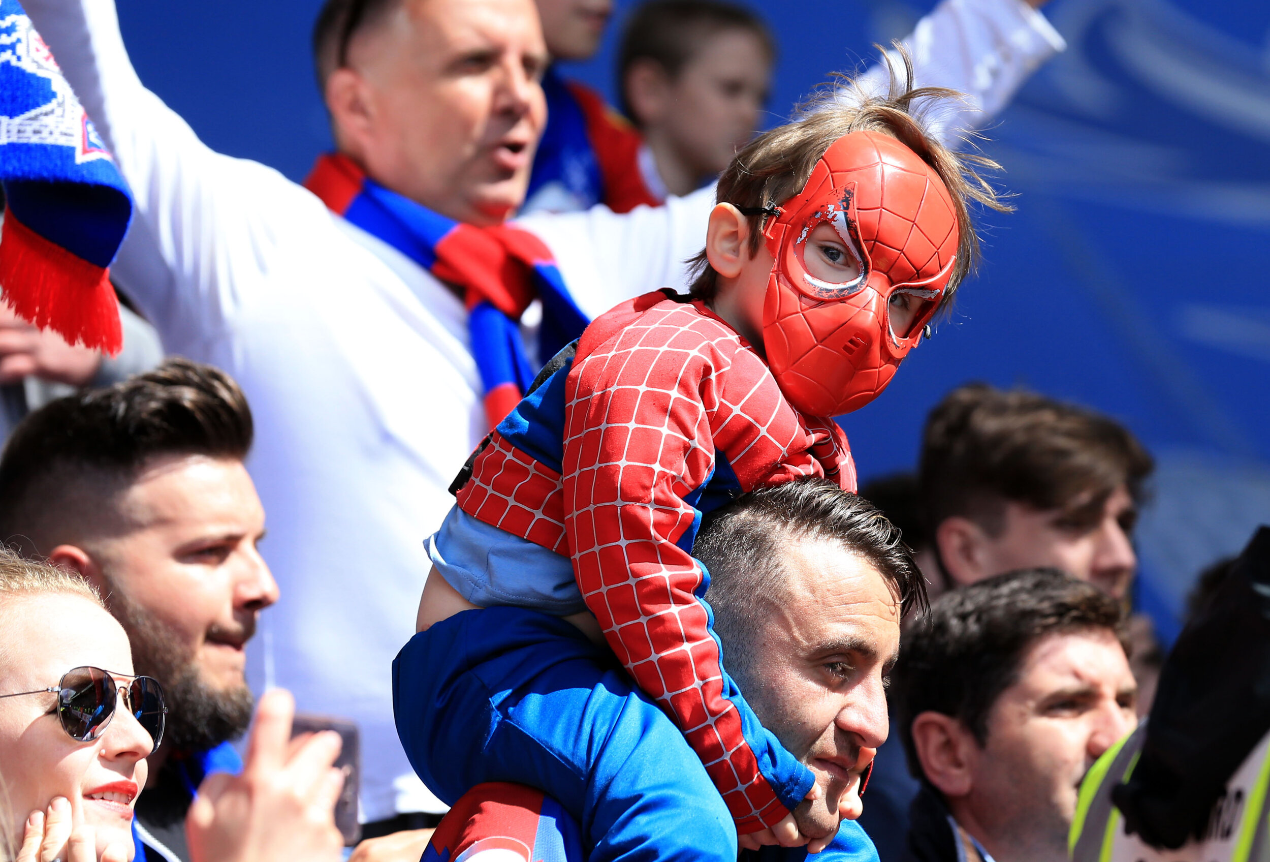 Child in Spider-Man costume on adult's shoulders at a sports event.