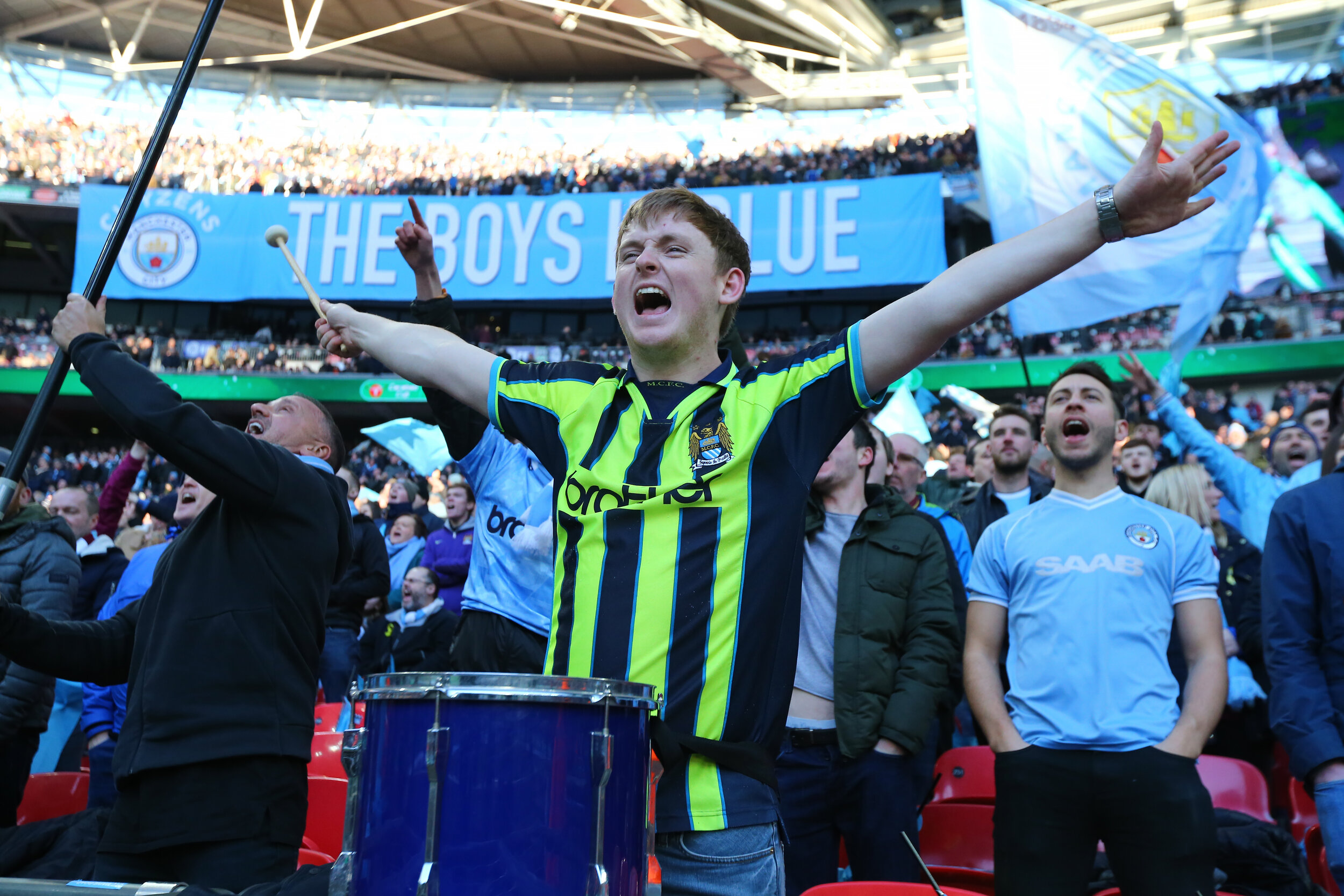 A group of enthusiastic soccer fans wearing blue and green jerseys cheer at a stadium. A prominent banner in the background reads "THE BOYS IN BLUE." One fan in the foreground is playing a large blue drum, while others raise their arms in excitement.