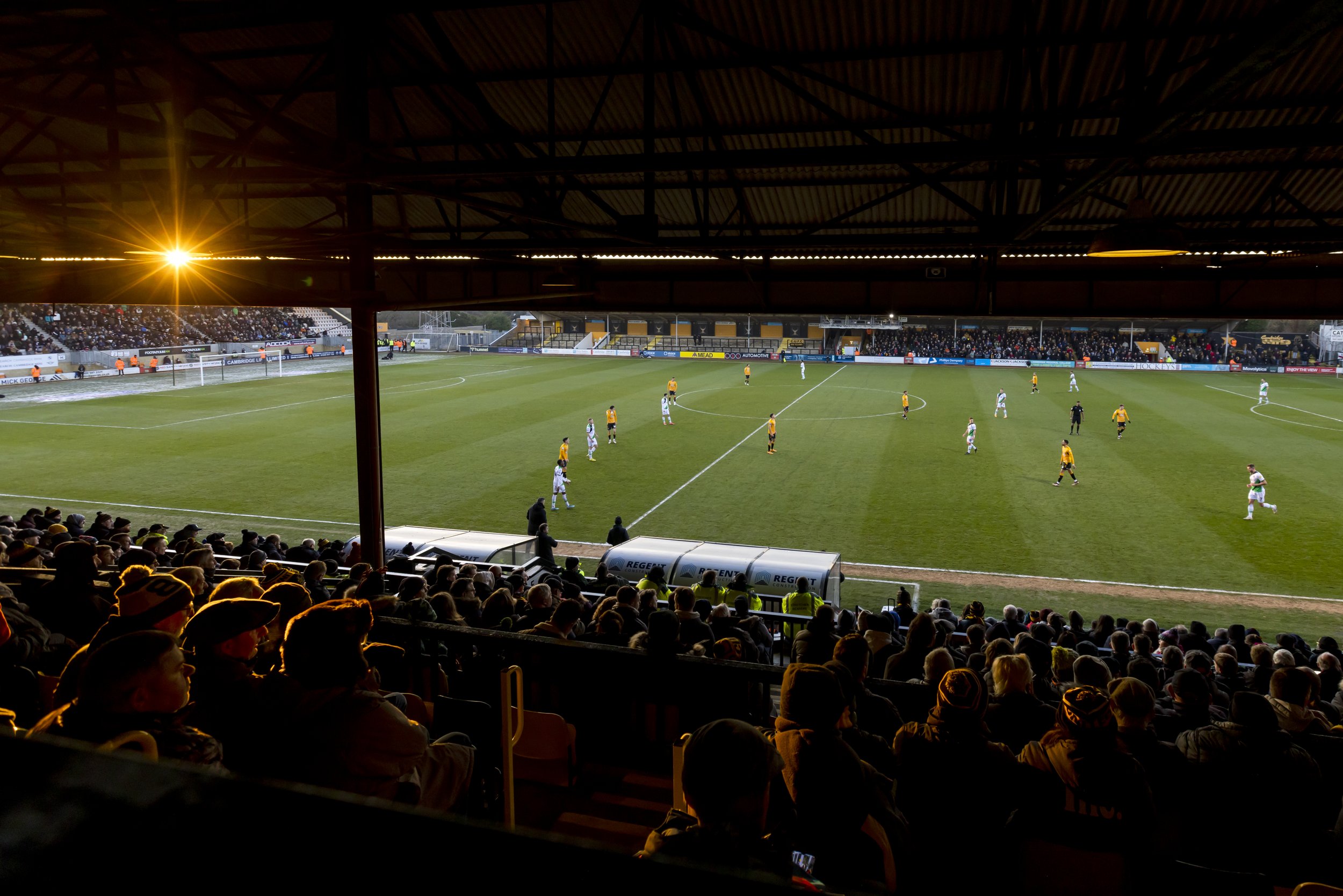 Soccer match in a crowded stadium with overhead roof at sunset.