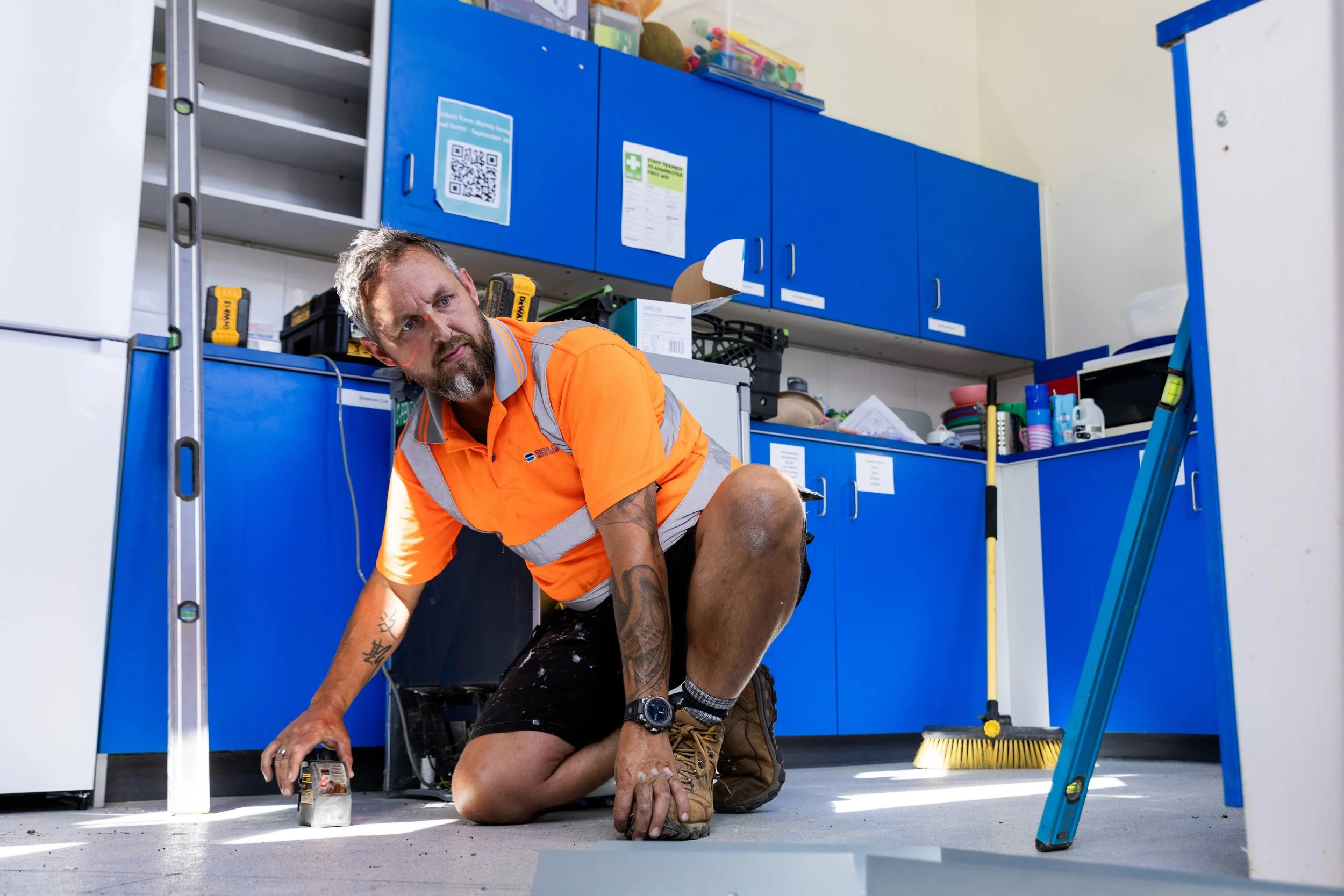 A man working on the floor of a workshop or storage room with blue cabinets, a level tool, a yellow broom, and various objects on the countertop, wearing an orange safety shirt and black shorts.