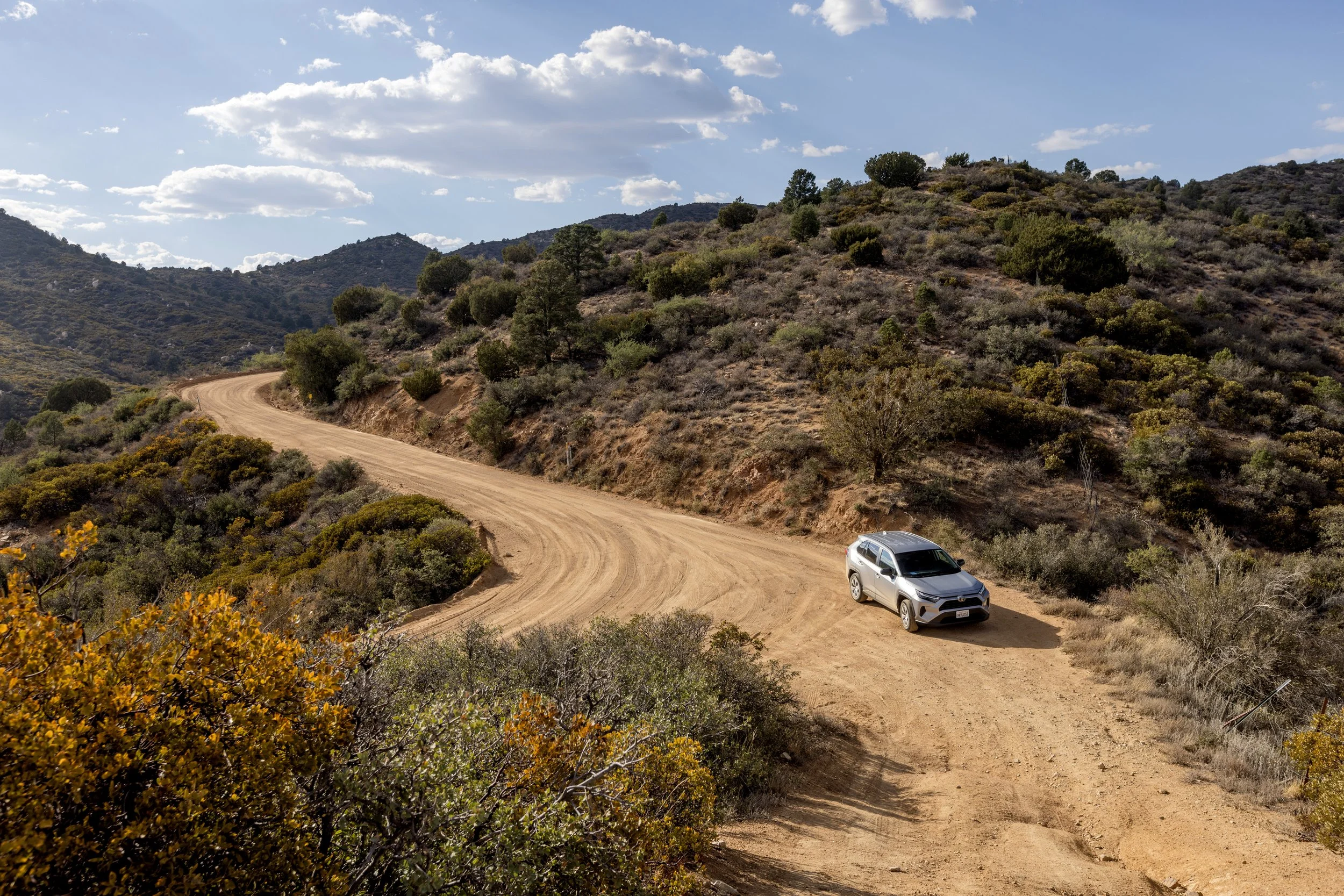 A silver SUV parked on a dirt winding mountain road surrounded by dry shrubbery and hills under a partly cloudy sky.