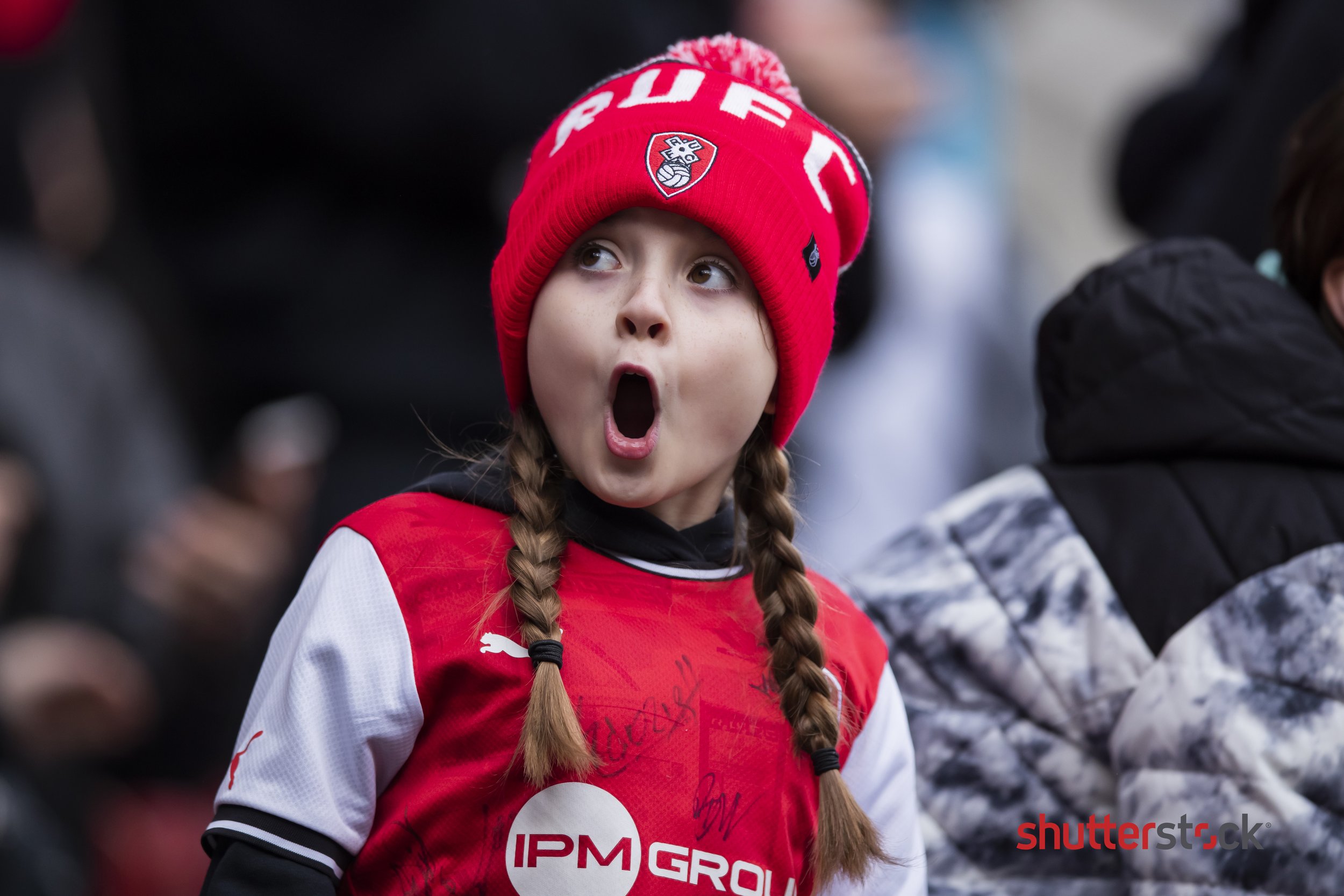 A young girl wearing a red beanie with "RUFC" and a football jersey, with an excited expression on her face at a sports event.