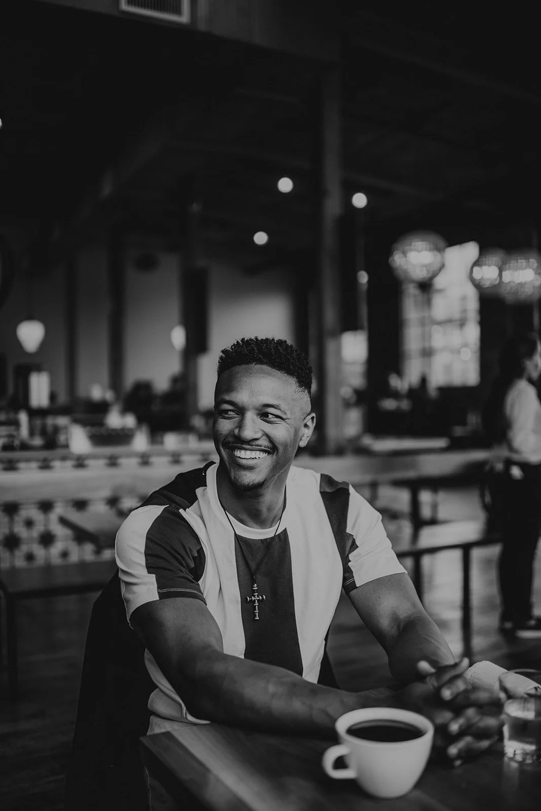 A smiling man sitting at a table in a cafe with a cup of coffee, in black and white.