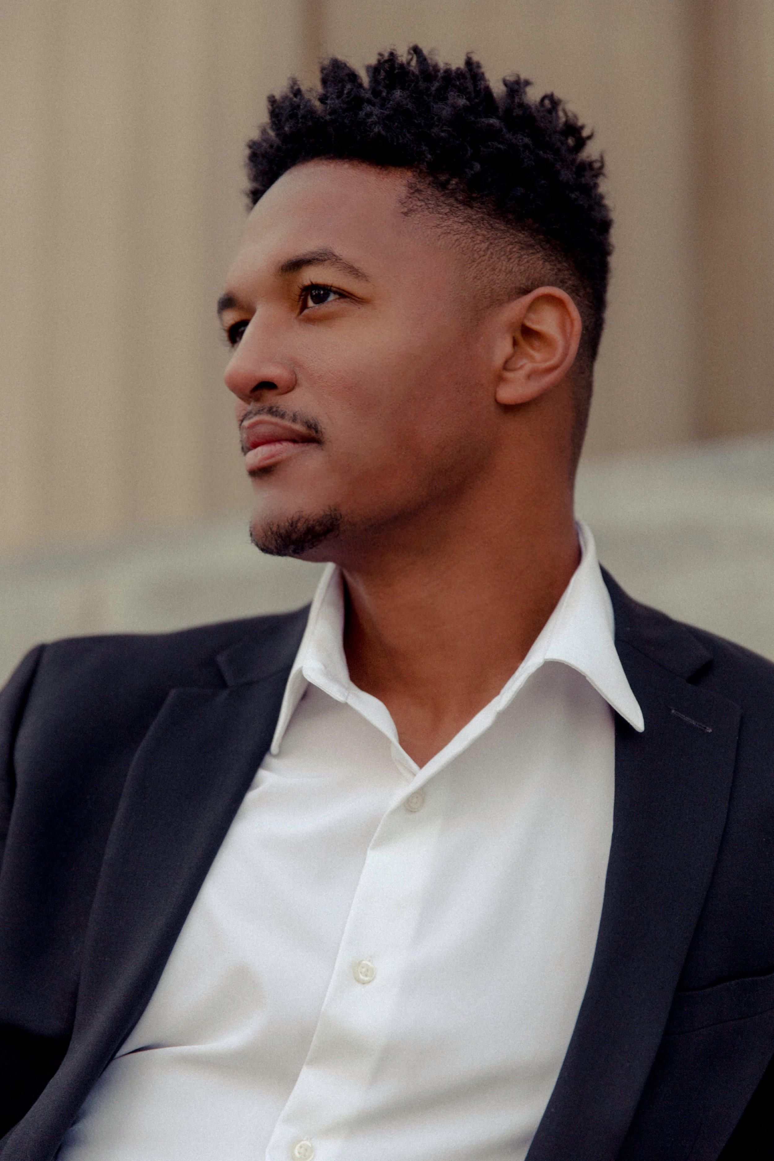 A young man in a black suit and white shirt gazes to the side, with a thoughtful expression. The background is blurred.