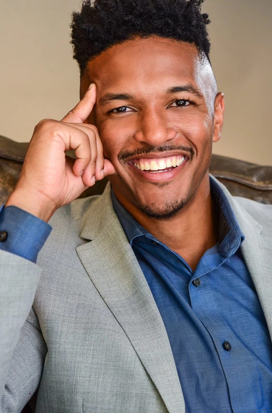 A smiling man in a gray suit and blue shirt sitting on a couch, with his hand resting on his temple.
