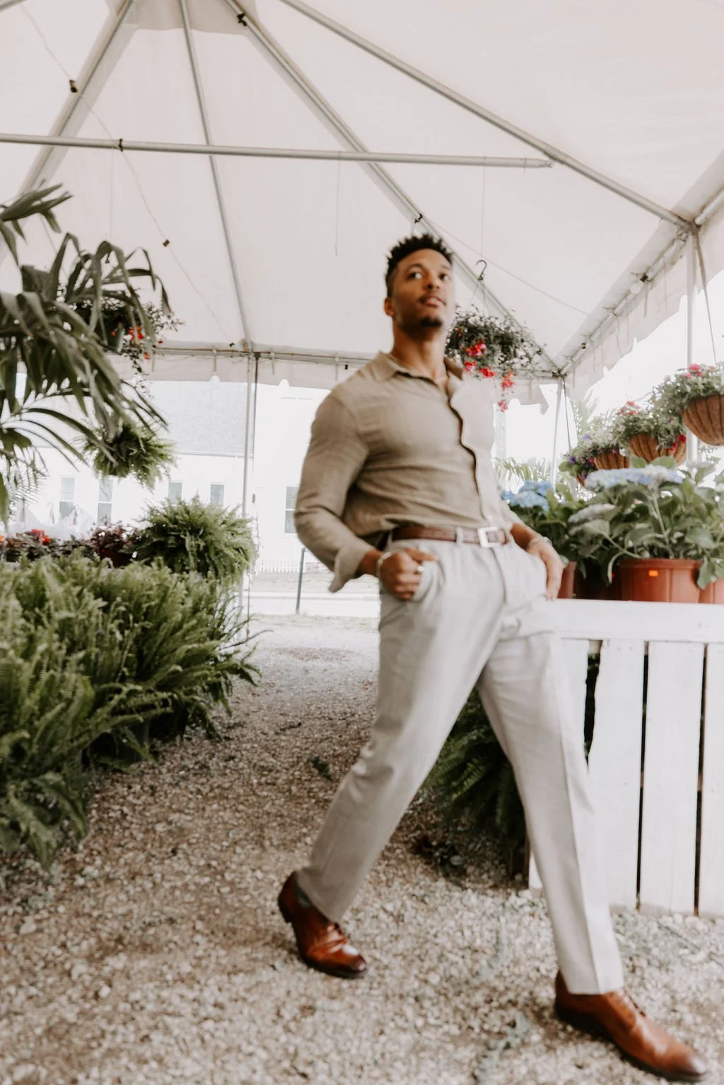 A young man in beige pants and a light brown shirt standing inside a greenhouse filled with potted plants and flowers.