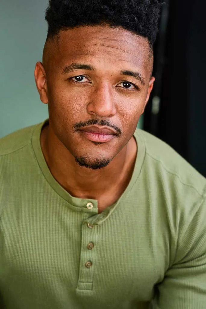 Close-up of a young man with short curly hair, a slight mustache and beard, wearing a green henley shirt, looking directly at the camera with a neutral expression.