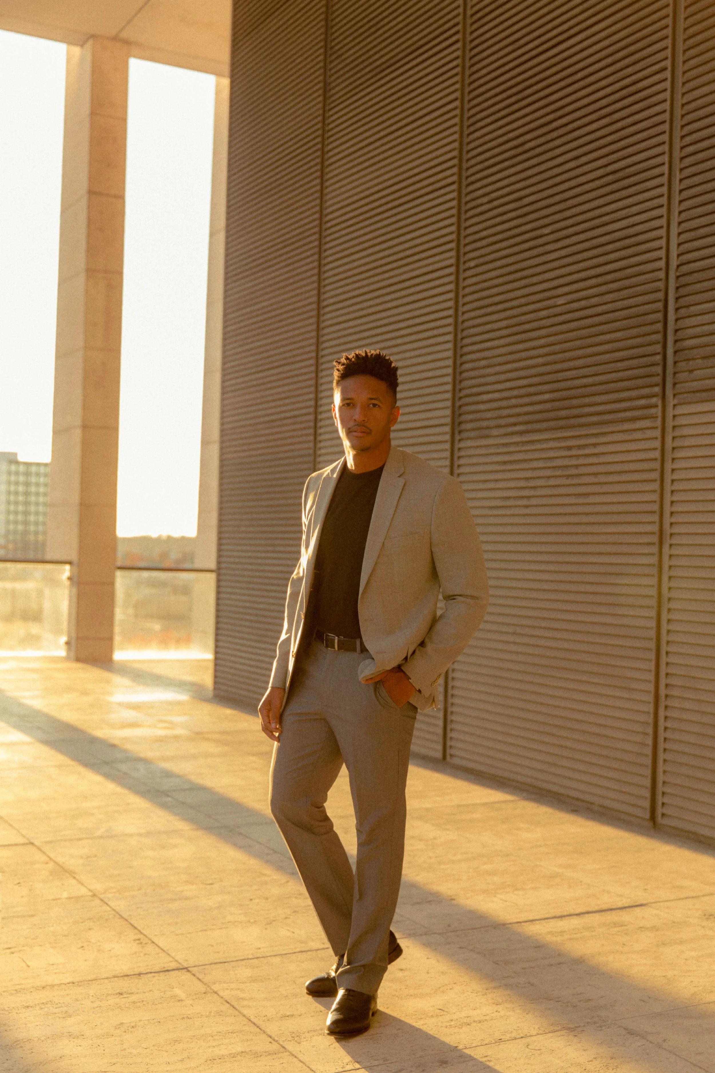 A man in a gray suit and black shirt standing on a balcony during sunset, with city buildings in the background.