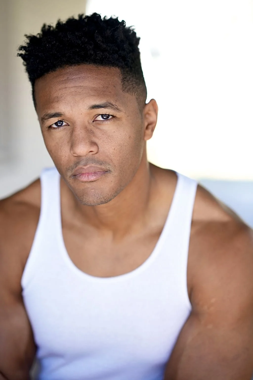 Close-up of a young man with dark hair and a white tank top, looking directly at the camera.