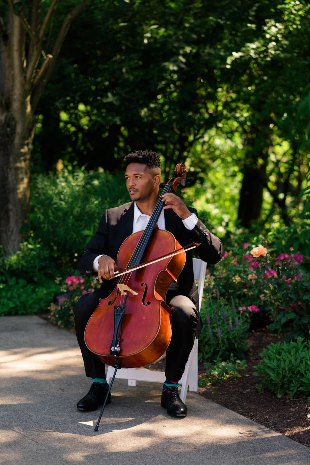 A man in a suit playing a cello outdoors with greenery and flowers in the background.