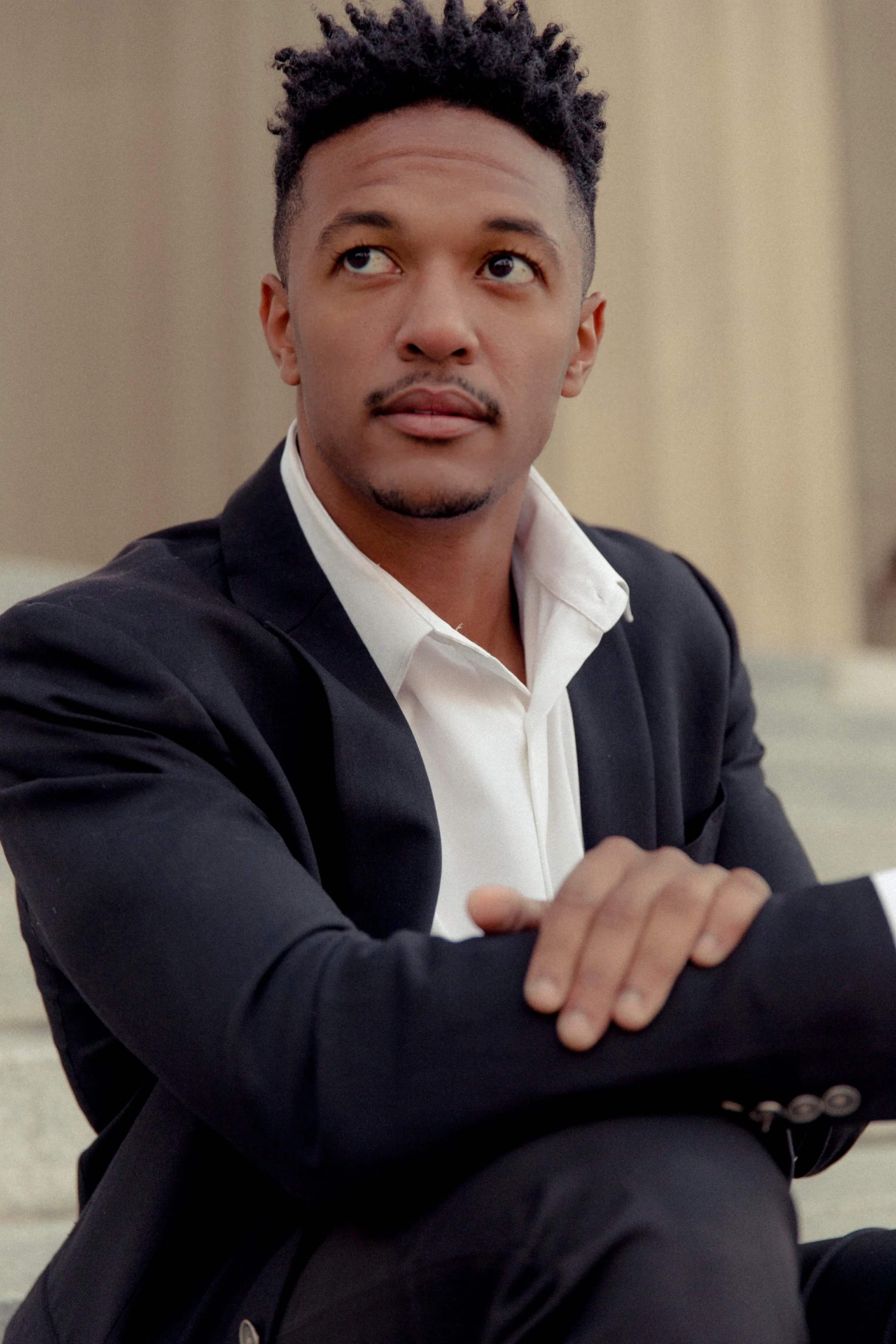 A young man in a black suit and white shirt sitting with arms crossed, looking to the side, with a neutral background.