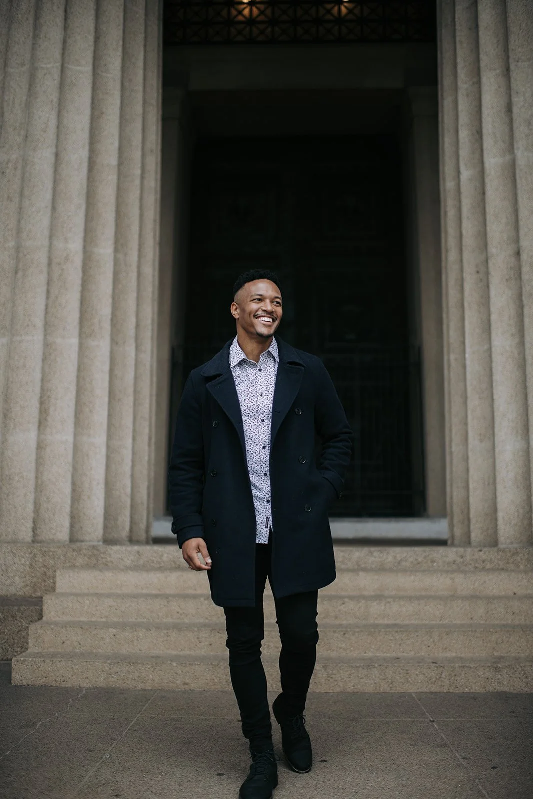 A smiling man in a patterned shirt and black coat walking down steps in front of a building with large columns.