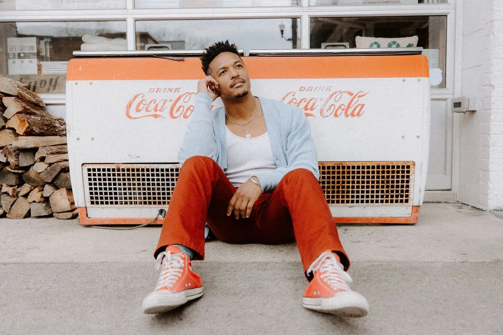 A young man sitting on the ground in front of a vintage Coca-Cola cooler, wearing red pants, red sneakers, a light blue jacket, and a white T-shirt, with a pile of firewood to his left.