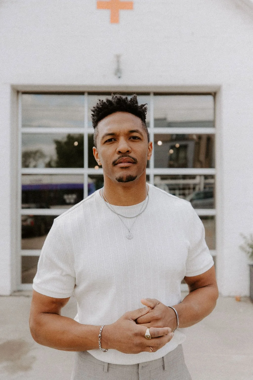 A man with short, curly black hair, a goatee, and rings on his fingers, wearing a white t-shirt, stands outside in front of a large glass window, with buildings and trees visible in the reflection.