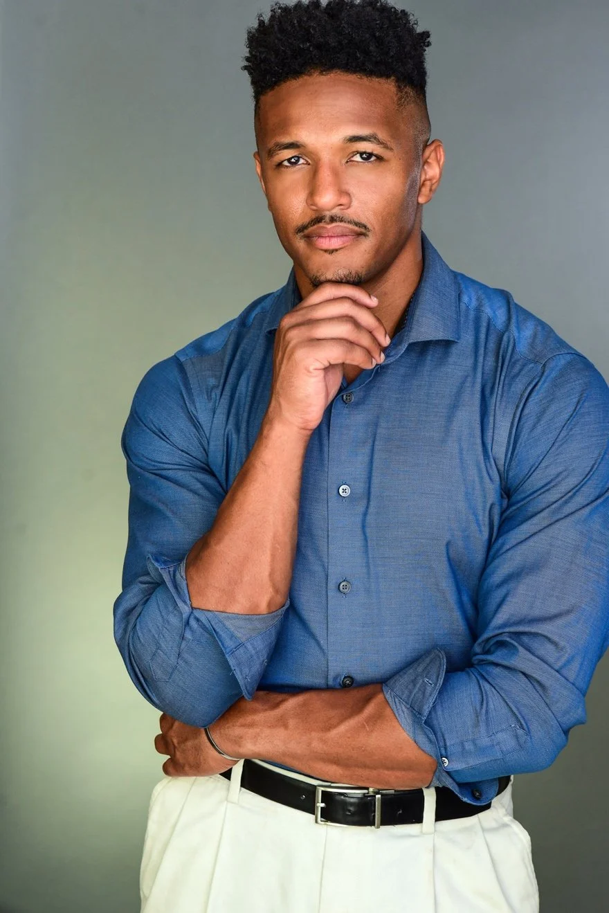 A young man in a blue dress shirt with rolled-up sleeves, beige pants, and a black belt, posing with one hand touching his chin and the other crossed over his abdomen, against a neutral background.