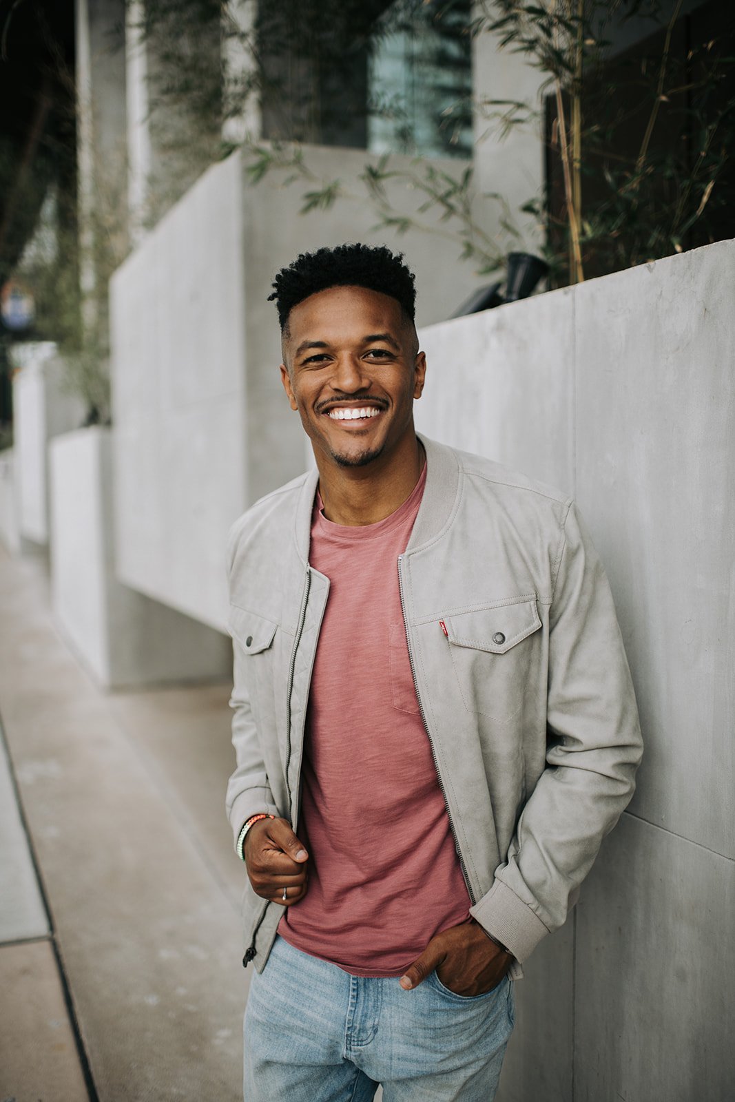 A man smiling and leaning casually against a white wall outdoors, dressed in a light-colored jacket, pink t-shirt, and jeans.