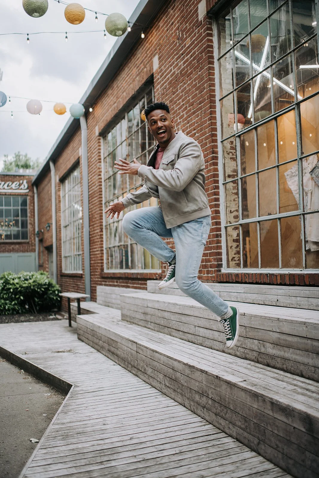 A young man is joyfully jumping off wooden steps outside a brick building with large windows, with colorful paper lanterns hanging above.